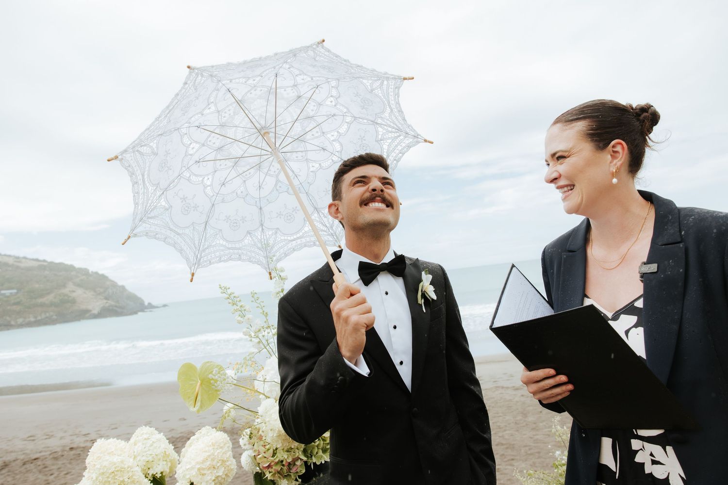 Wedding ceremony setup on a beach with white umbrellas and floral arrangements against the ocean backdrop.