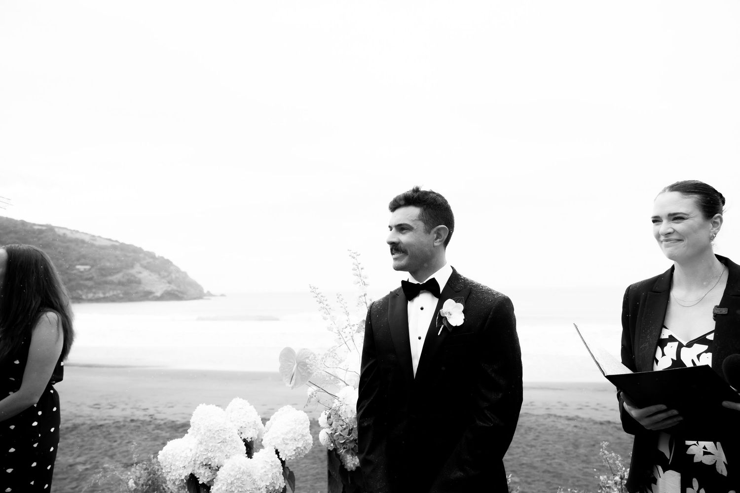 A black and white seaside wedding ceremony photo with coastal cliffs in the background.