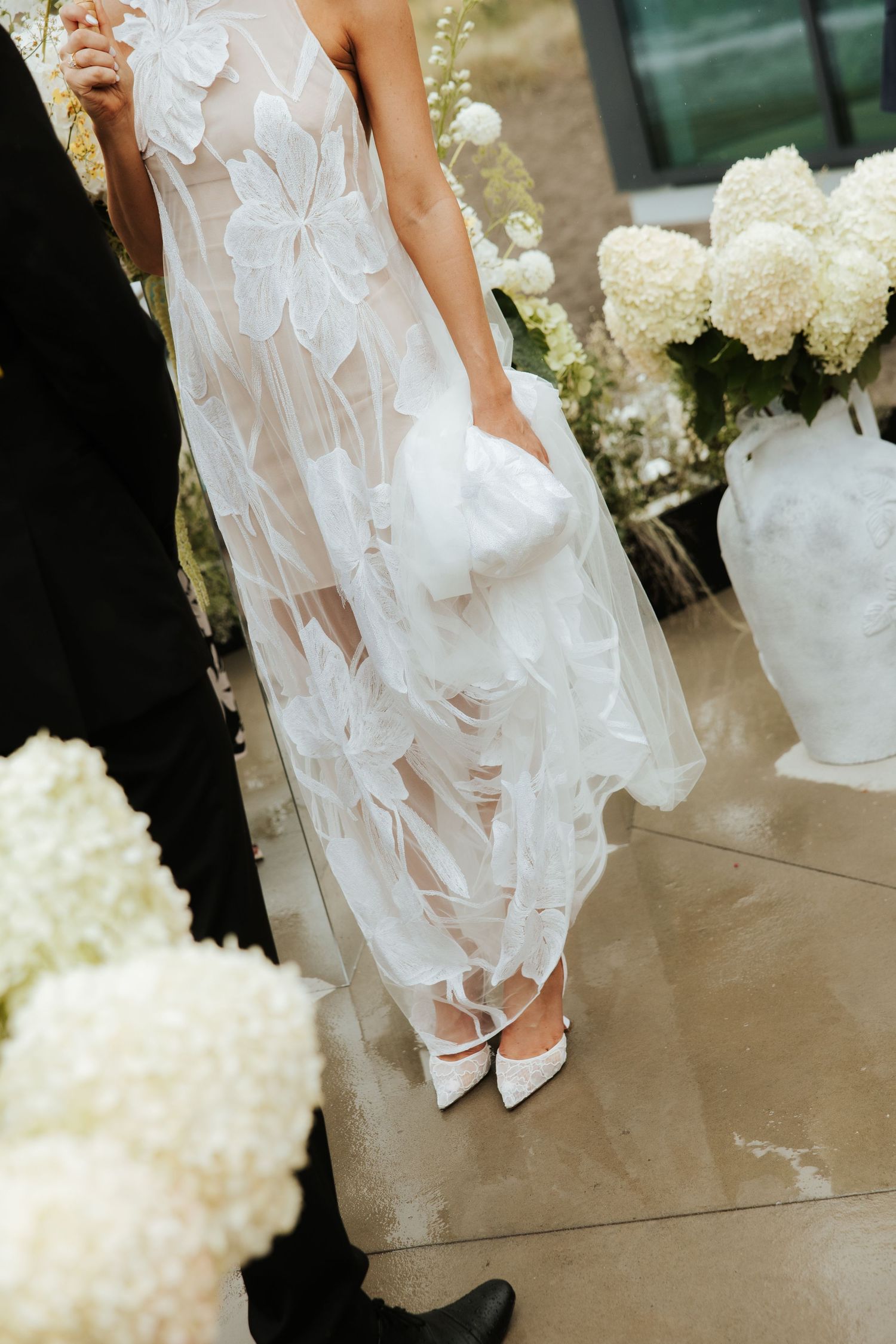 Detail of floral lace wedding dress and white heels surrounded by hydrangeas.