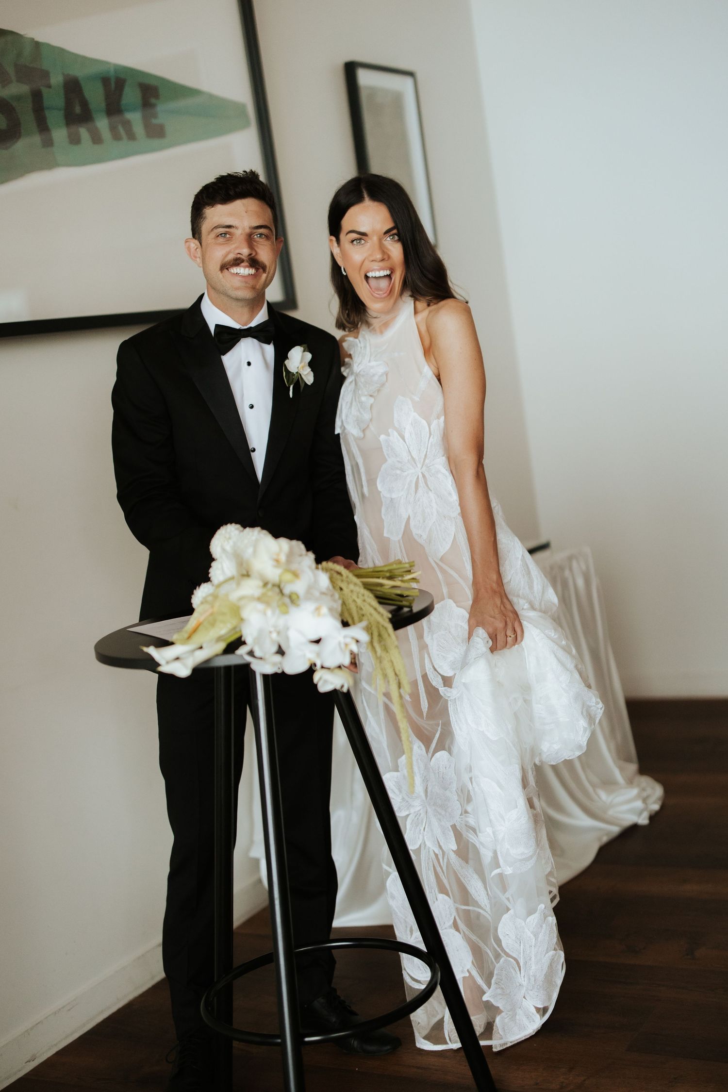A newlywed couple poses with white flowers in formal attire against a minimalist interior.