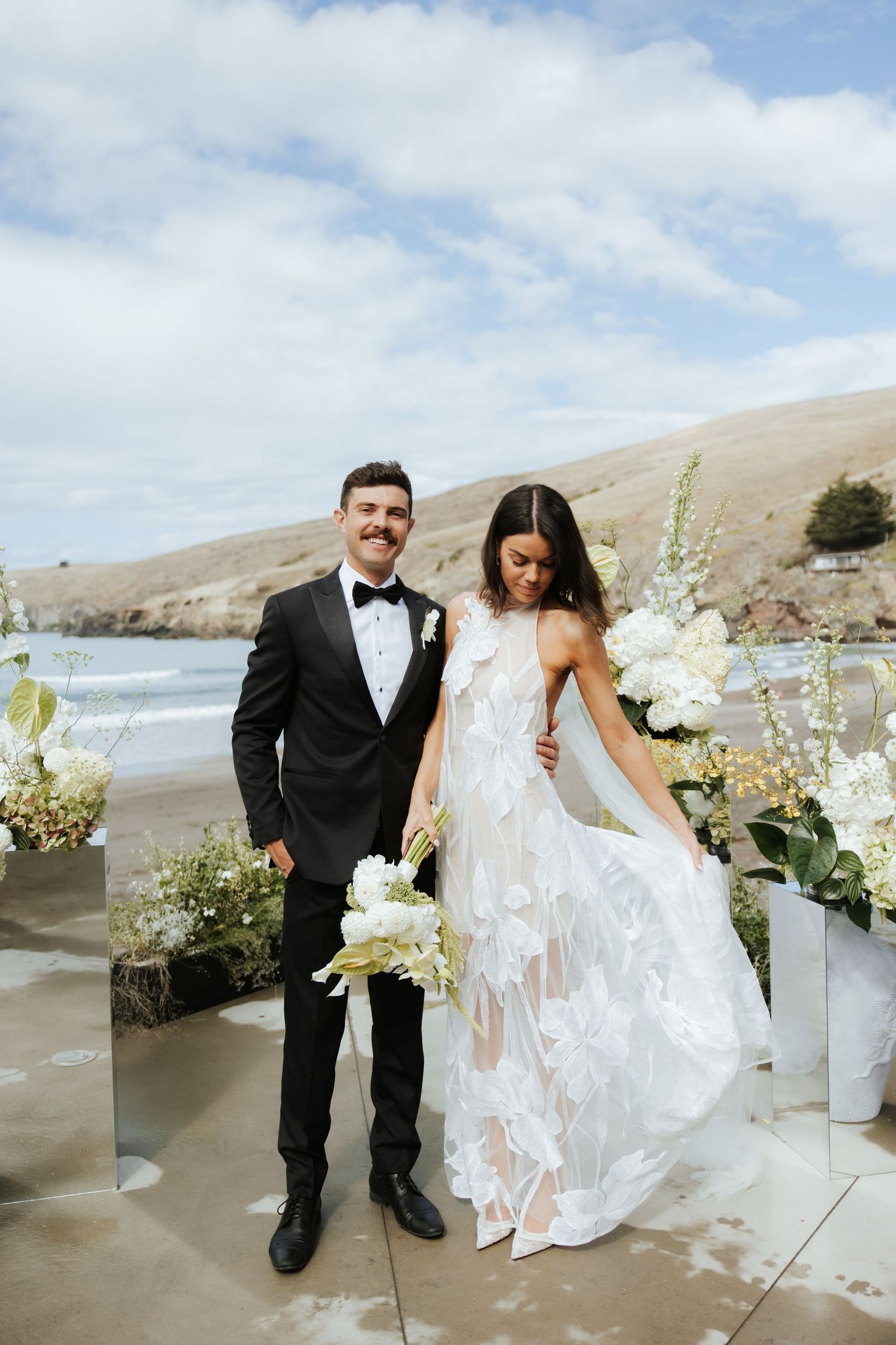 Wedding couple poses by elegant white floral arrangements on a scenic beach.