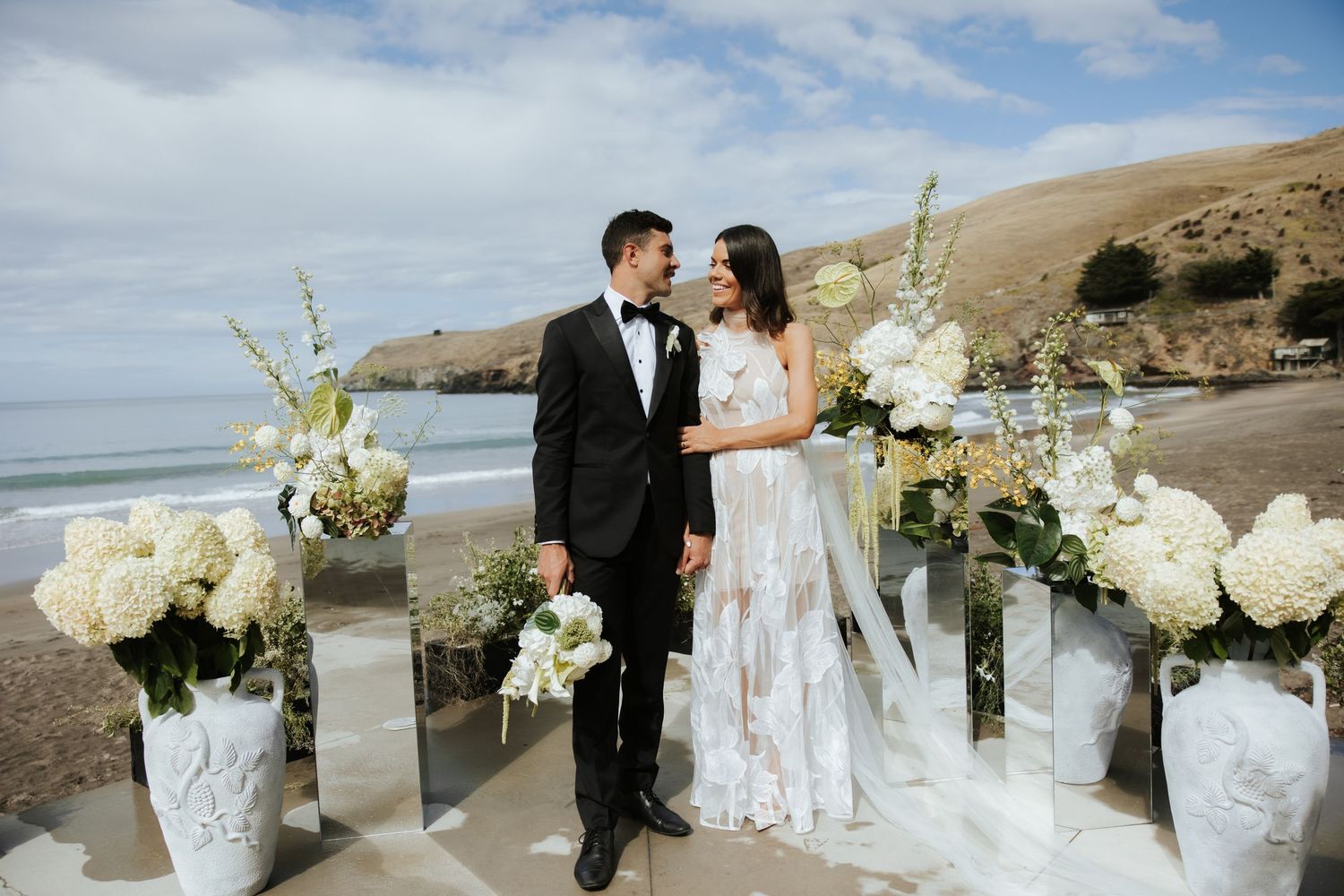 Elegant white floral arrangements in vases decorate a coastal wedding ceremony site.