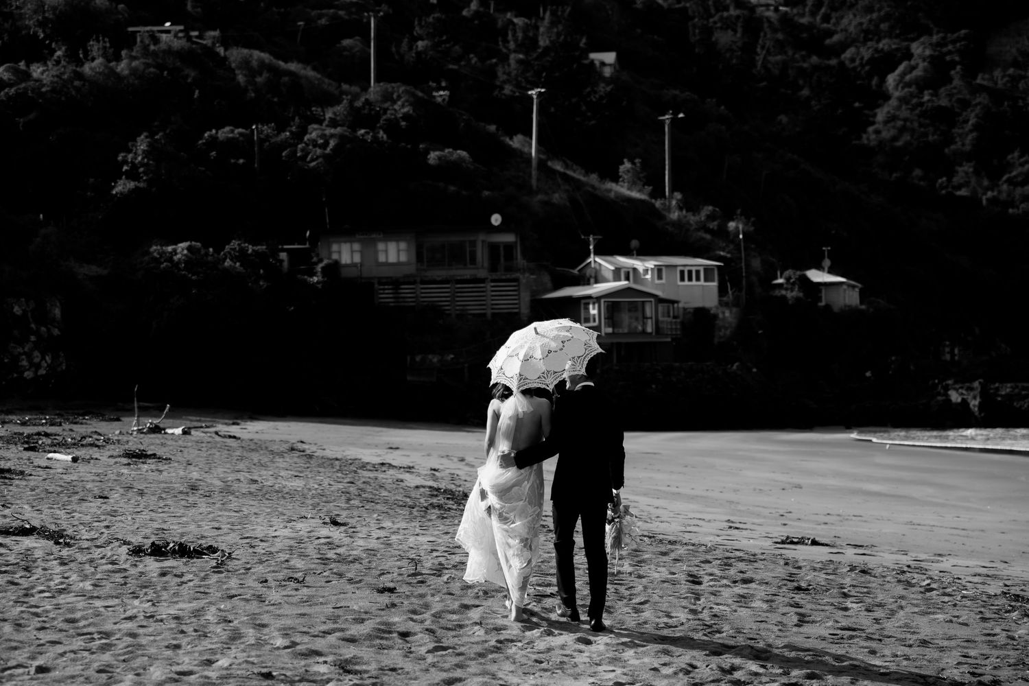 A black and white photo of newlyweds walking together on a beach holding a parasol.