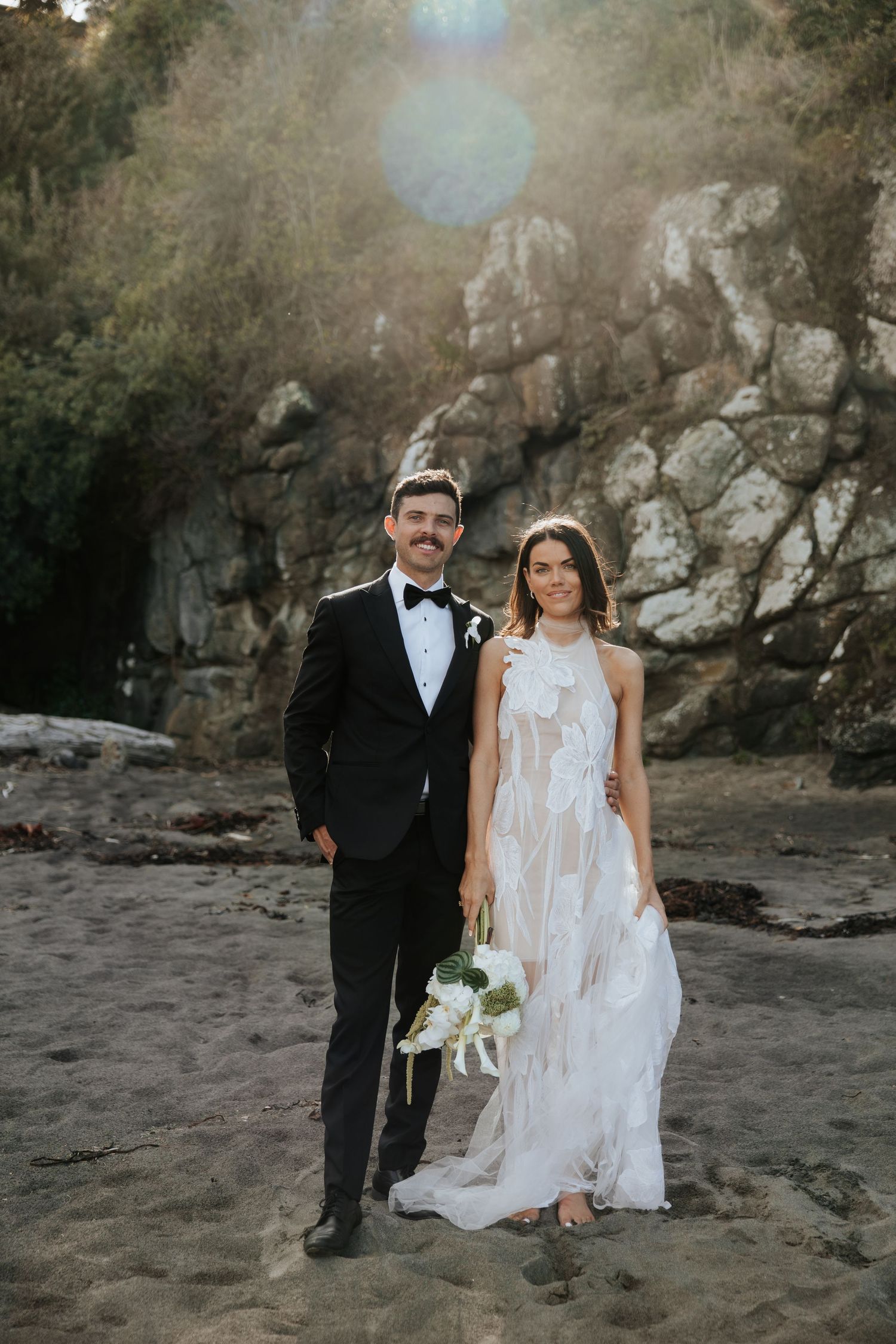 Couple in wedding attire poses against rocky beach cliffs and natural scenery.