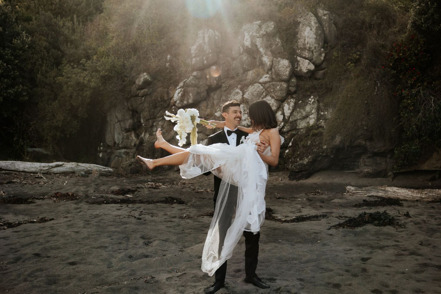 A dramatic wedding photo of a couple embracing on a beach with rocky cliffs in the background.
