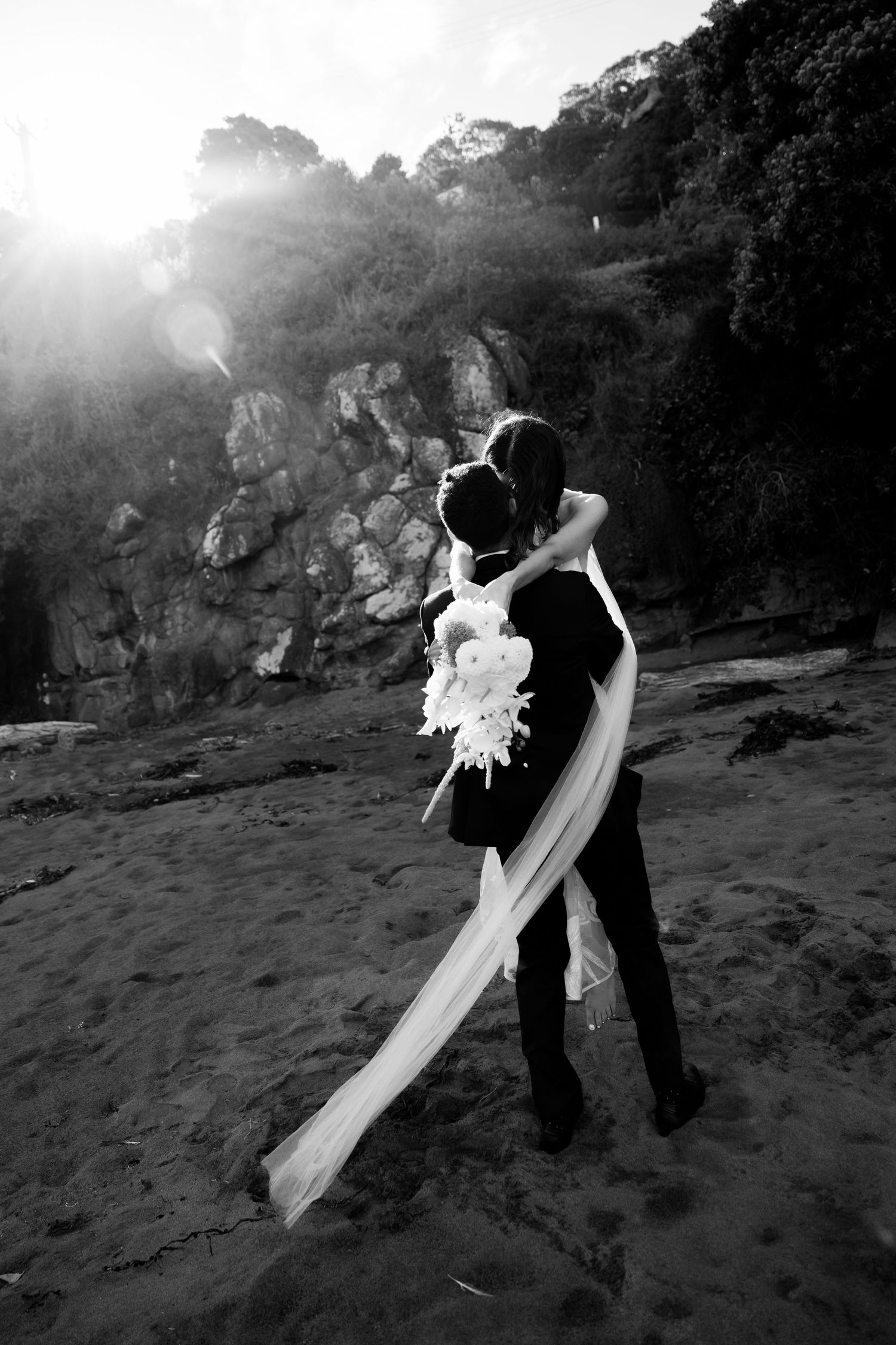 A black and white silhouette of a couple embracing on the beach with sunlight streaming through rocks.