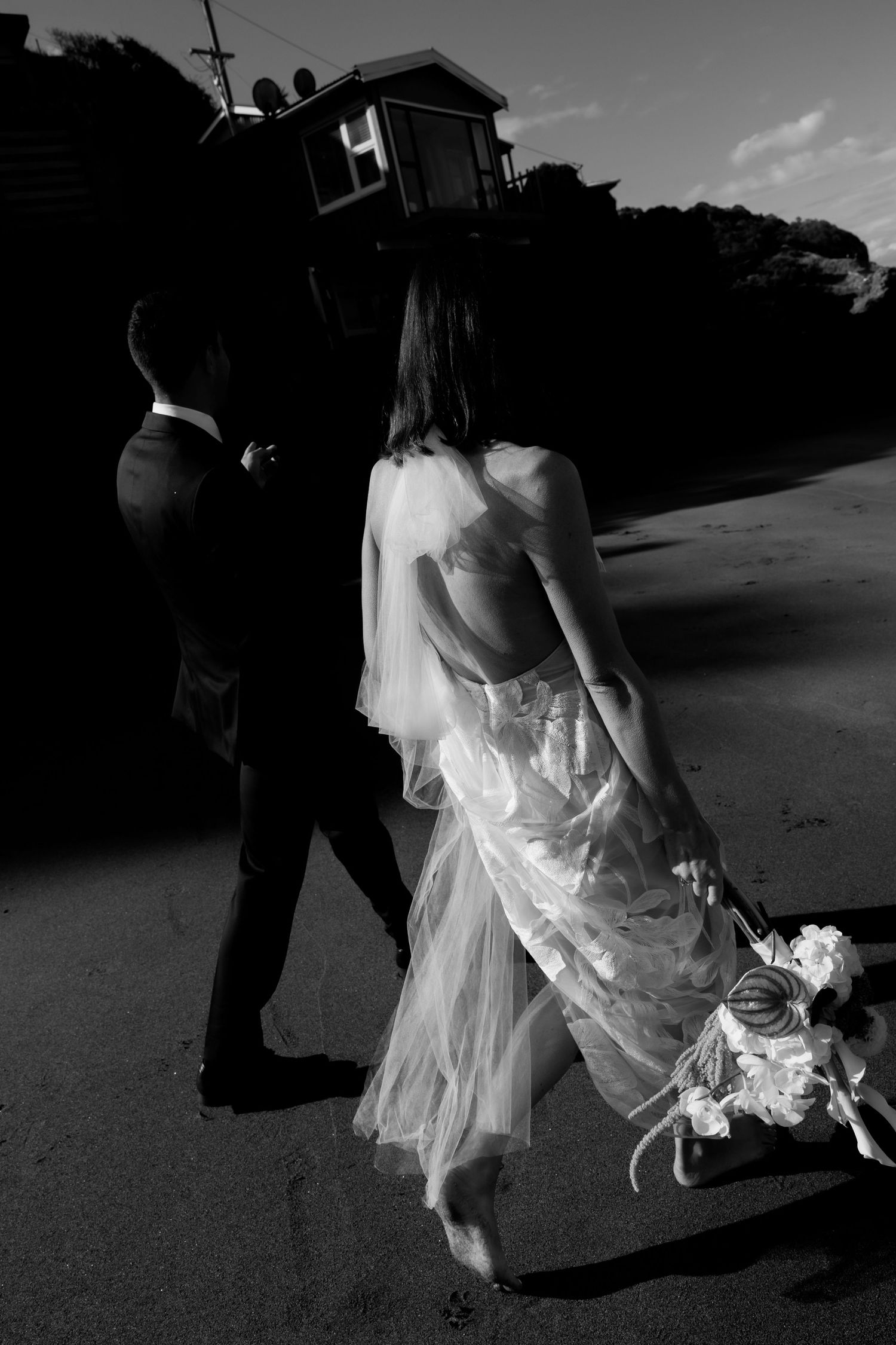 Black and white photo of newlyweds walking together, with bride holding bouquet against dramatic lighting.