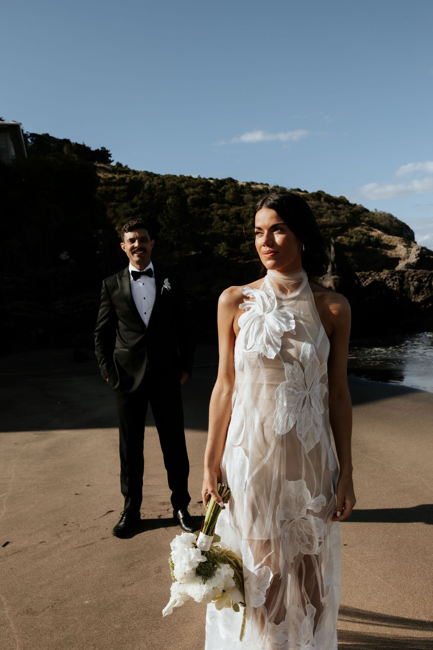 A couple poses on a beach for their wedding photos, with the bride in a white flowy dress holding a bouquet.