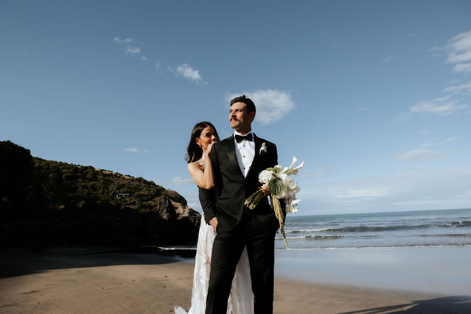 A wedding couple stands together on a sunny beach with cliffs and ocean waves in the background.