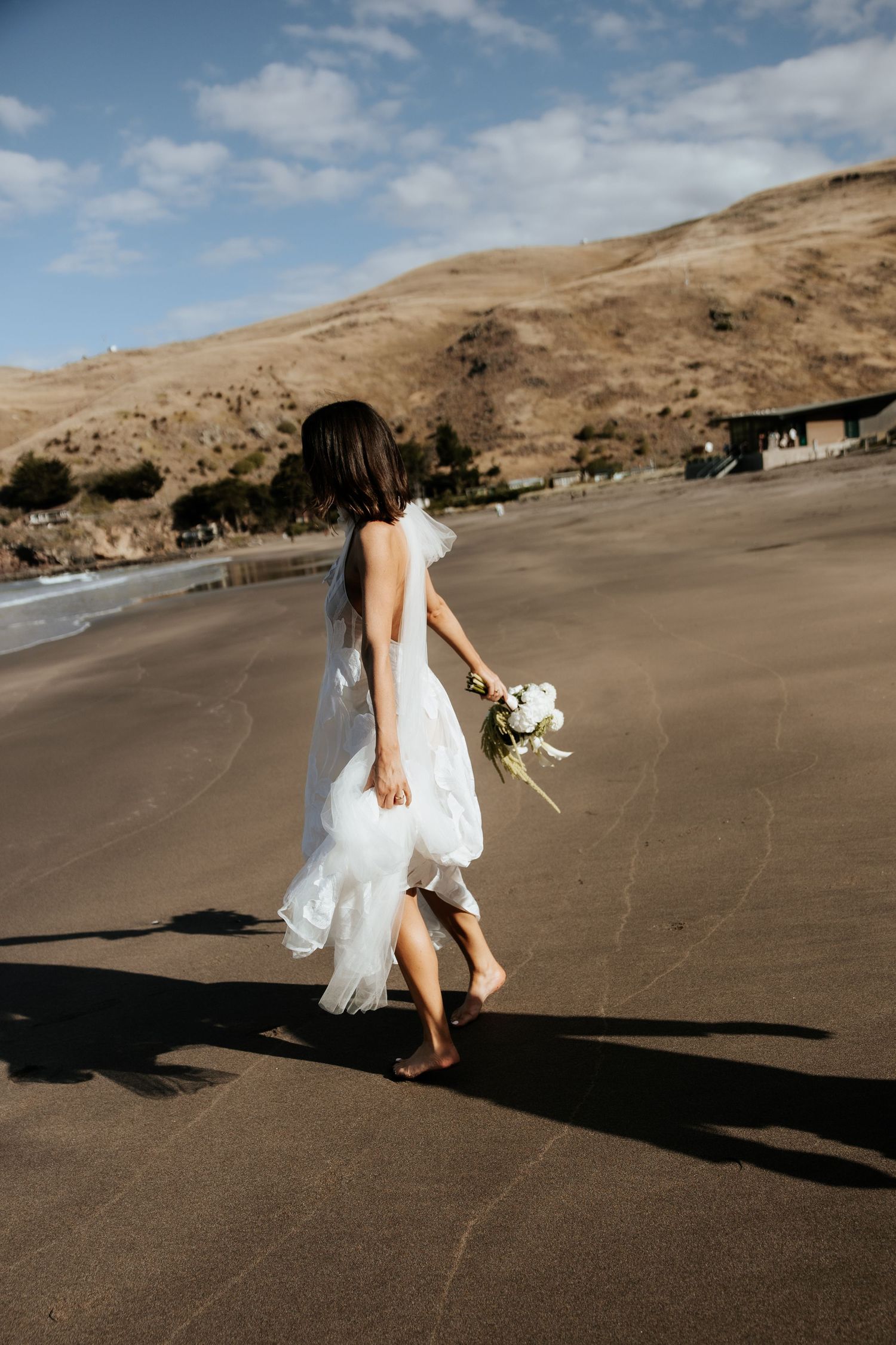 Bride in white dress walking barefoot on a black sand beach with coastal hills in background.