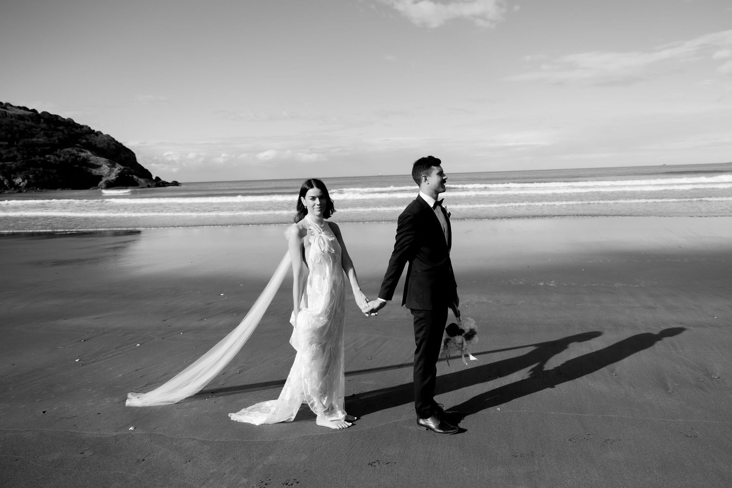 Wedding couple walking hand in hand on a scenic beach with long shadows cast on the sand.