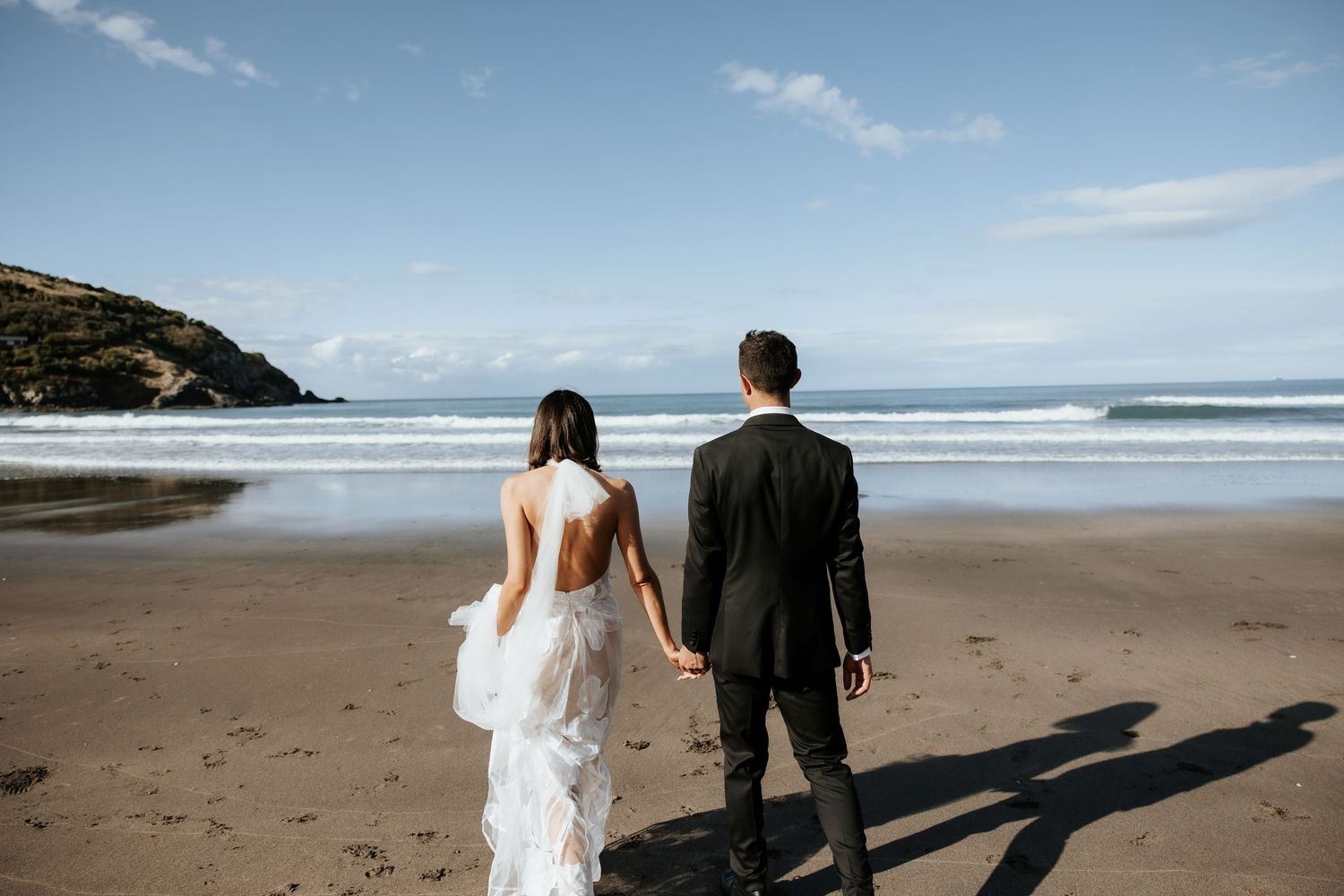 Couple walking along a scenic beach with waves and mountains in the distance.