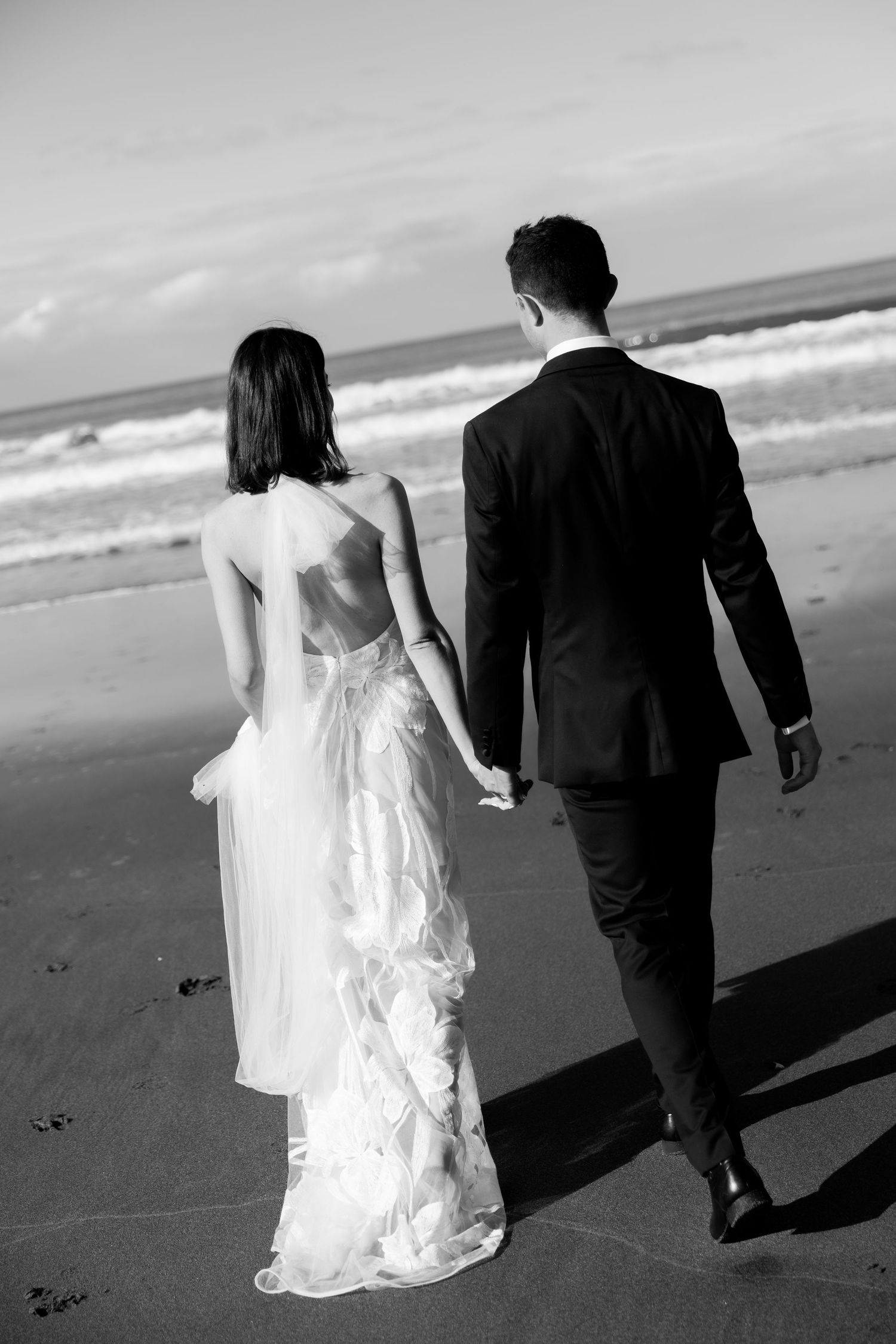 A couple in wedding attire walks hand in hand along a beach in black and white.