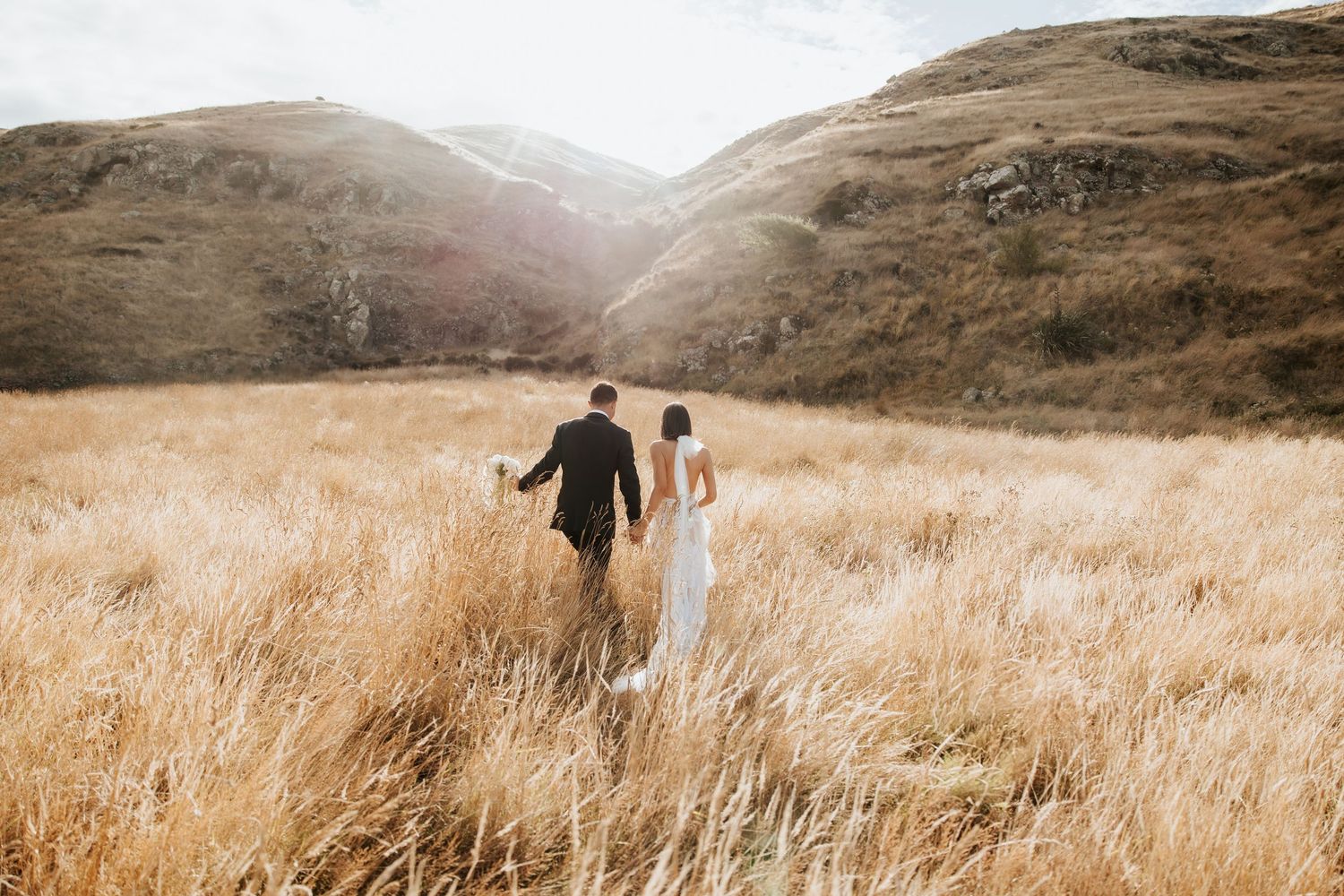 Couple walks through golden grass field towards mountains during sunset.