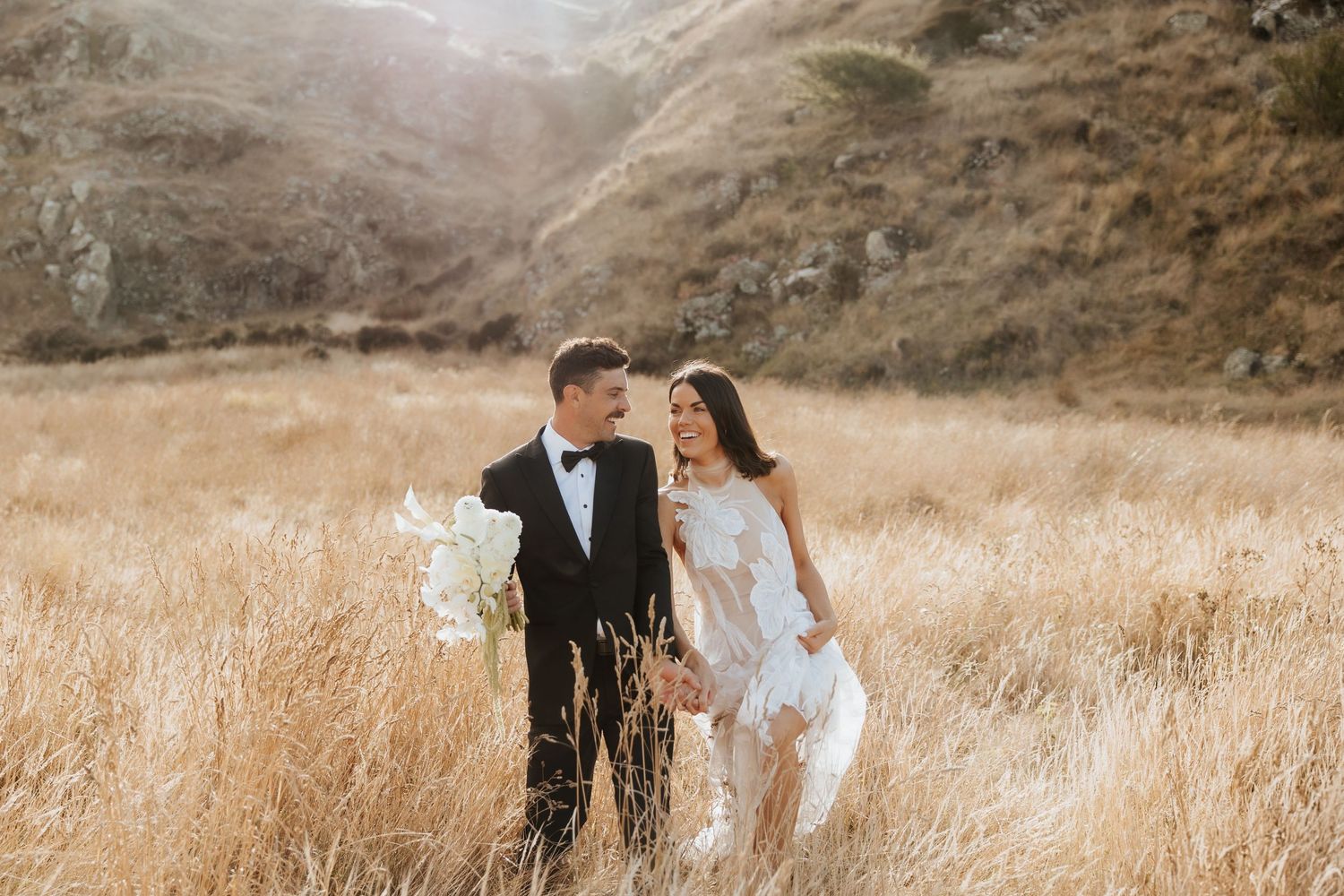 Wedding couple walking through golden grass field with mountain backdrop.