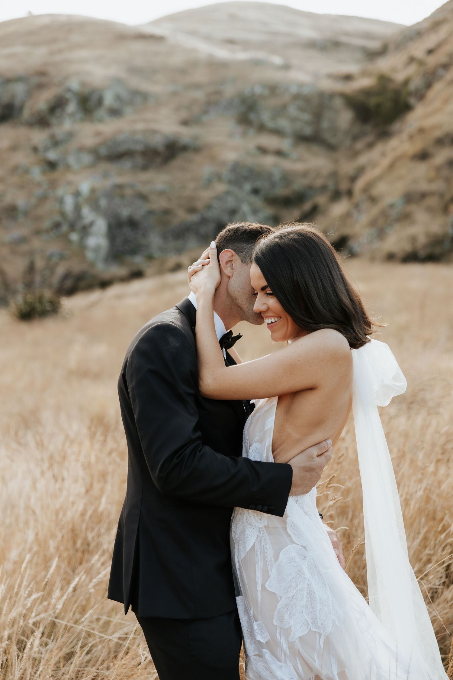 A romantic outdoor wedding scene with the couple embracing in golden grass against a mountainous backdrop.