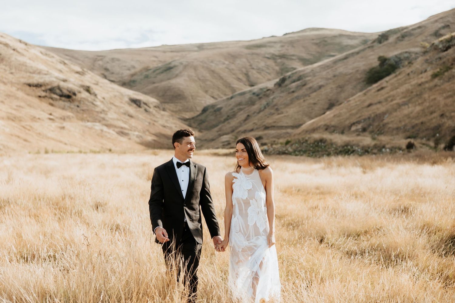A couple in formal wear walking through golden grass with mountains in the background.