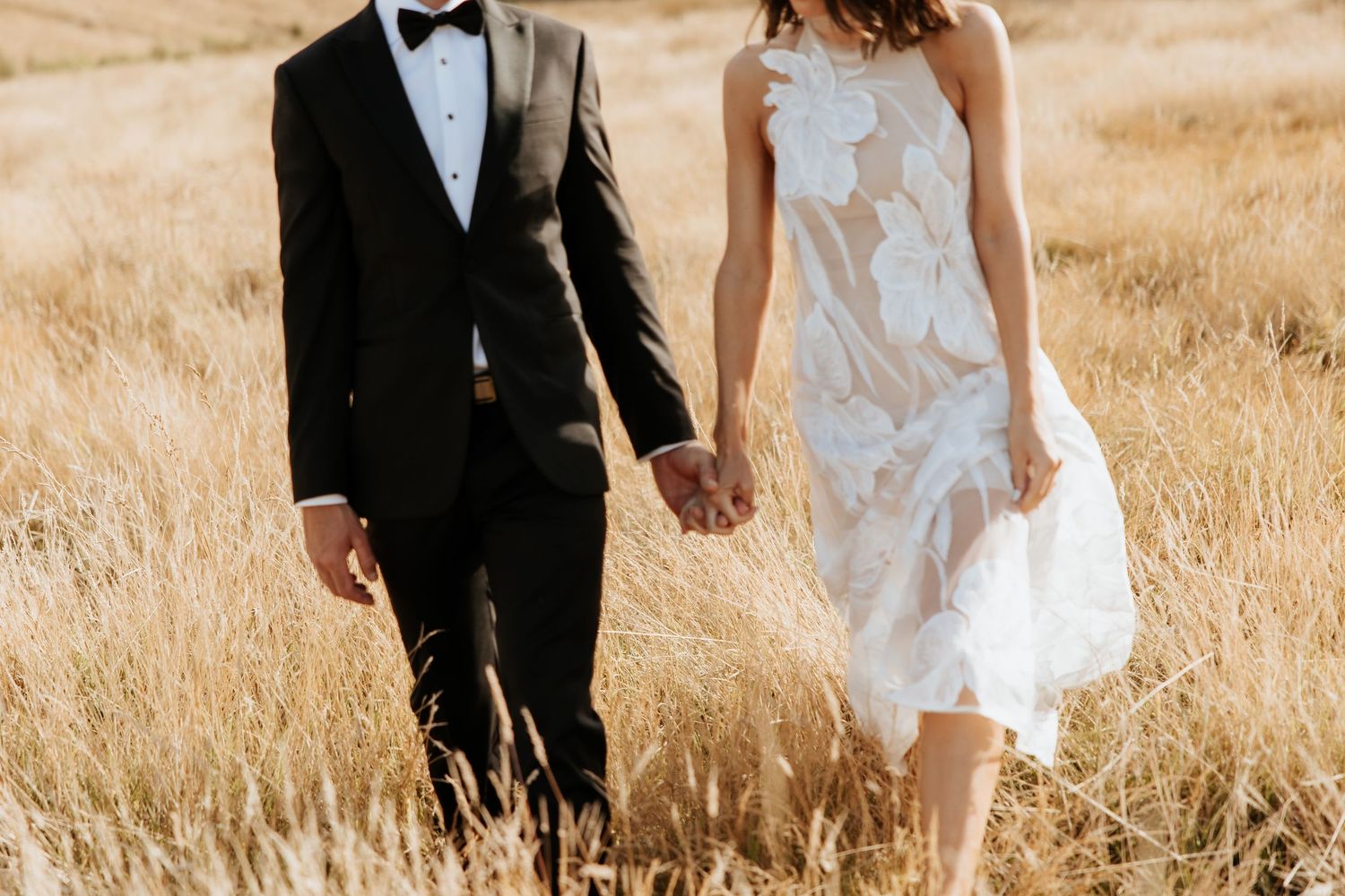 Couple in wedding attire walking hand in hand through golden grass field.