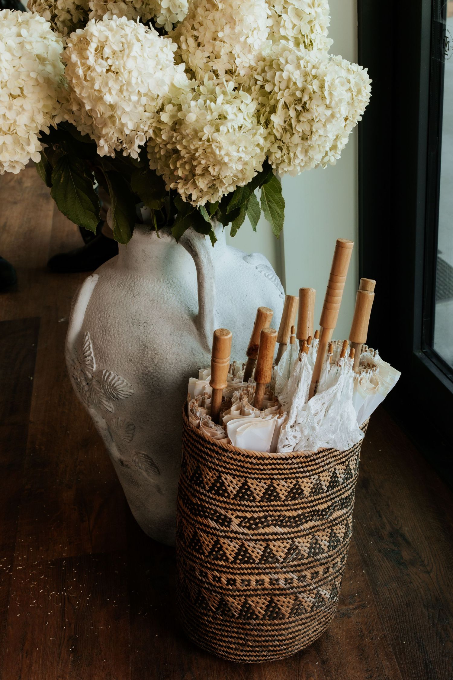 White hydrangea flowers arranged in a ceramic vase next to a decorative basket with cinnamon sticks on a wooden surface.