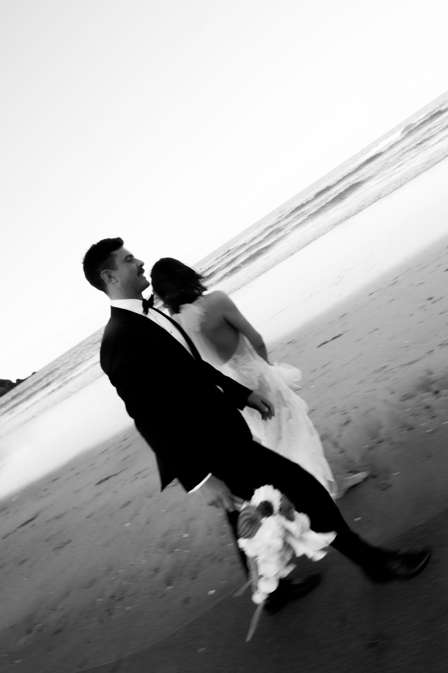 A black and white beach wedding photo shows a couple in formal attire dancing by the ocean.
