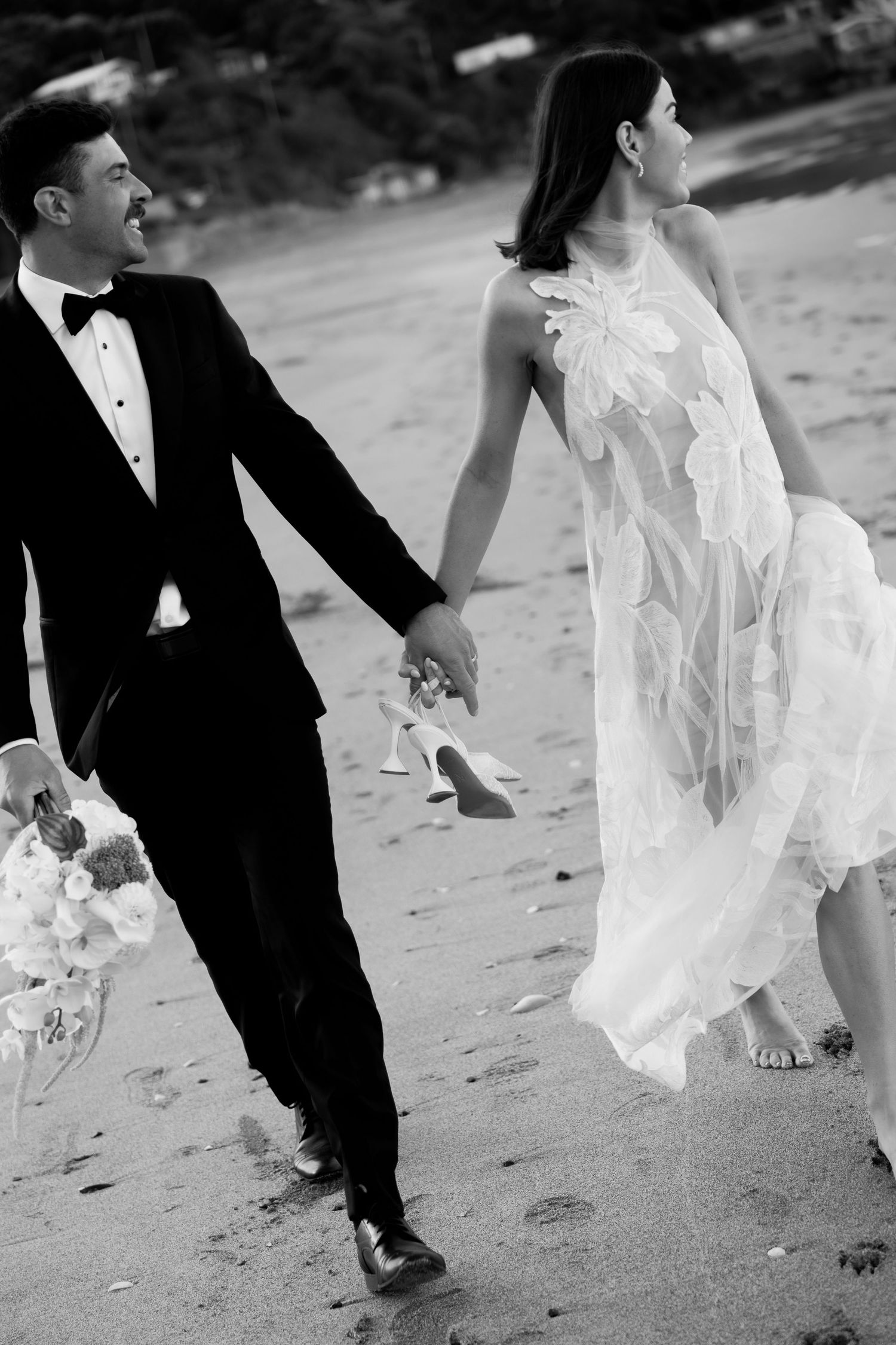 A romantic black and white photo of a couple in formal wear walking hand in hand on a beach.