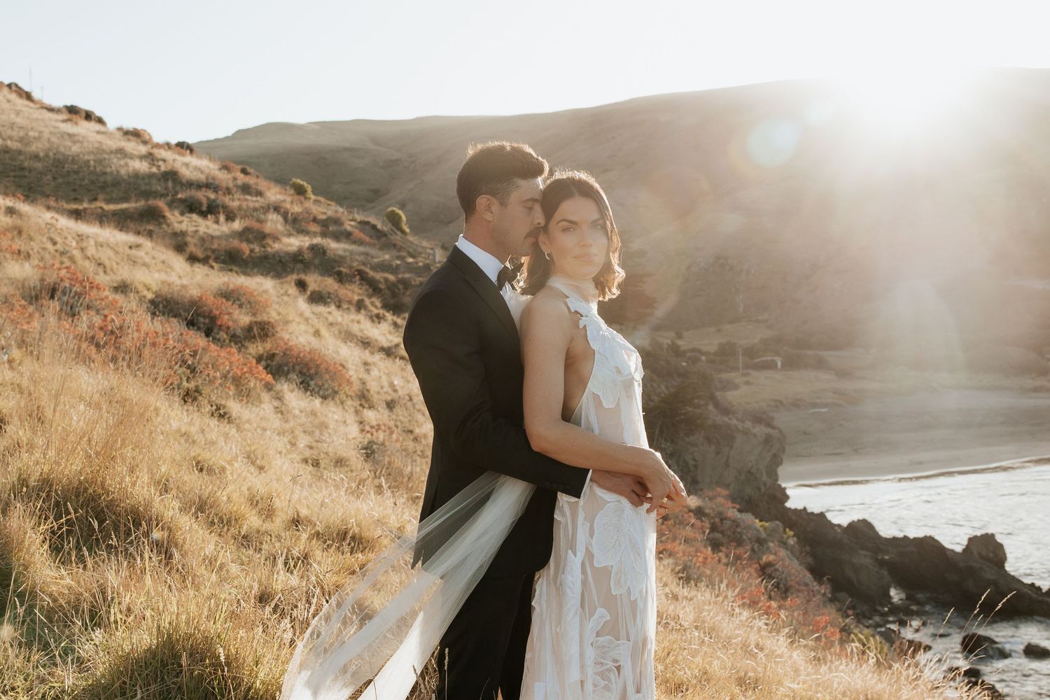 Wedding couple embraces on hillside overlooking coastal views at golden hour.