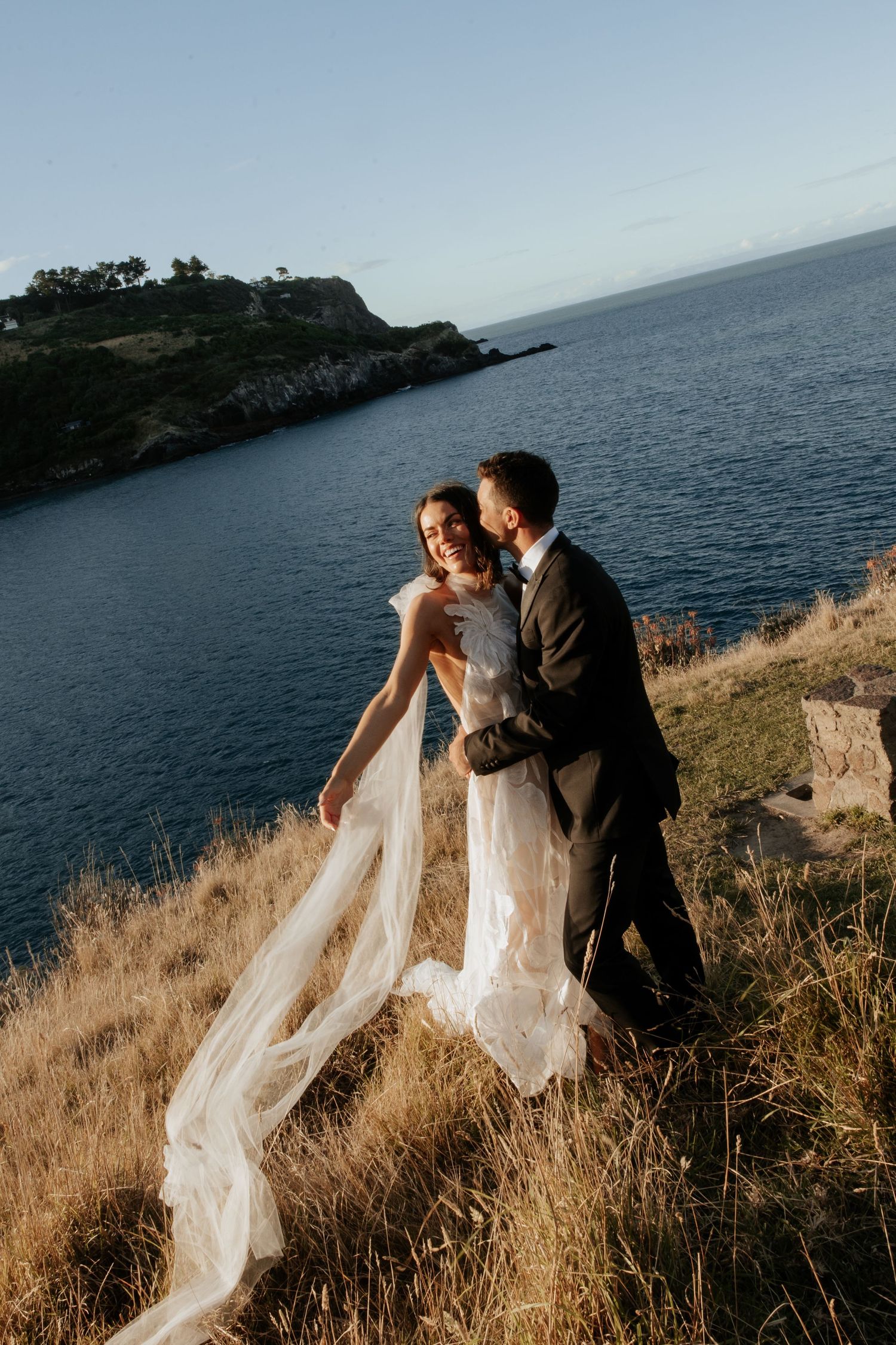 Romantic coastal wedding portrait against dramatic ocean cliffs at sunset.