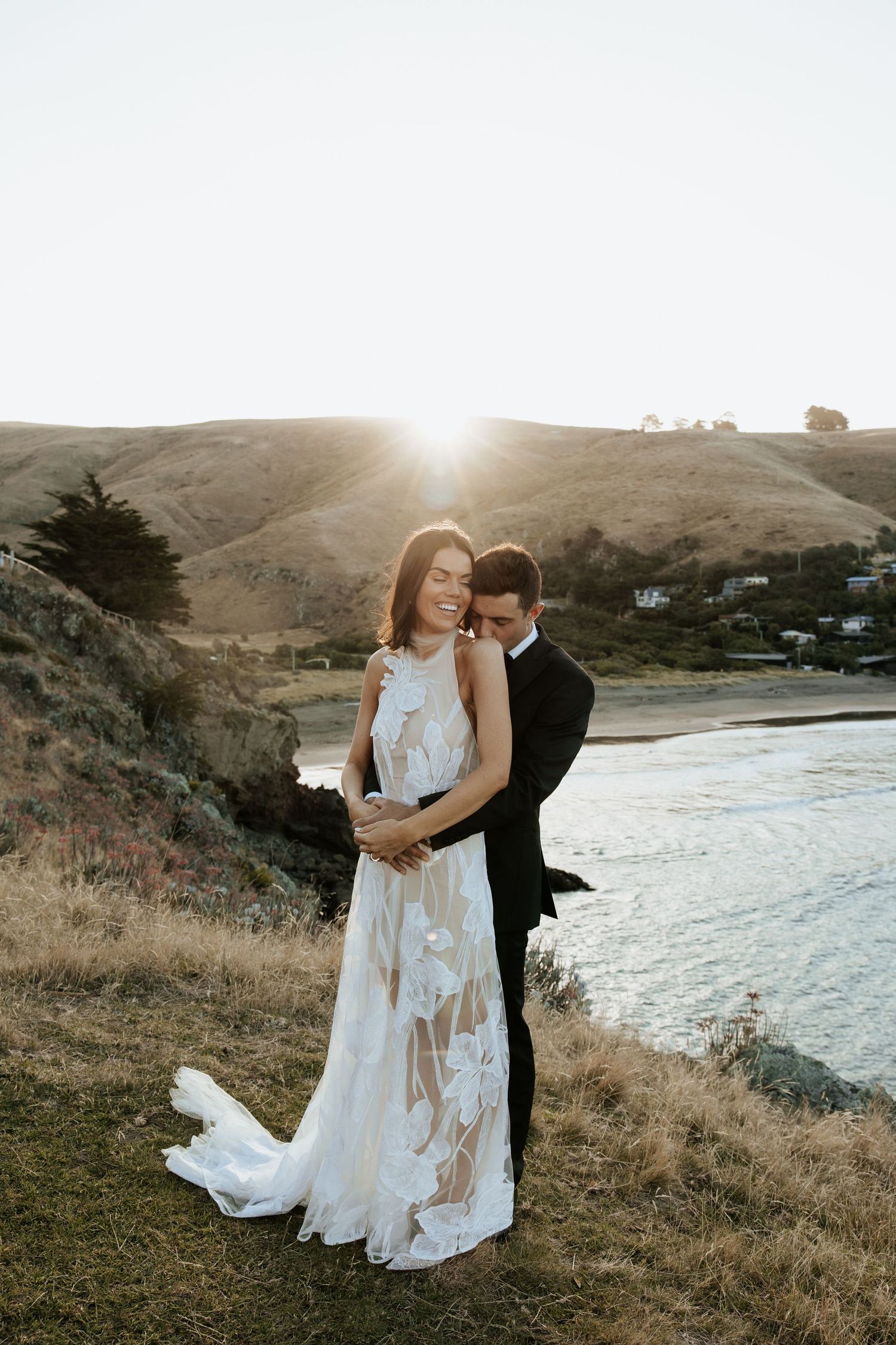 A couple embraces during sunset on a coastal clifftop in wedding attire.