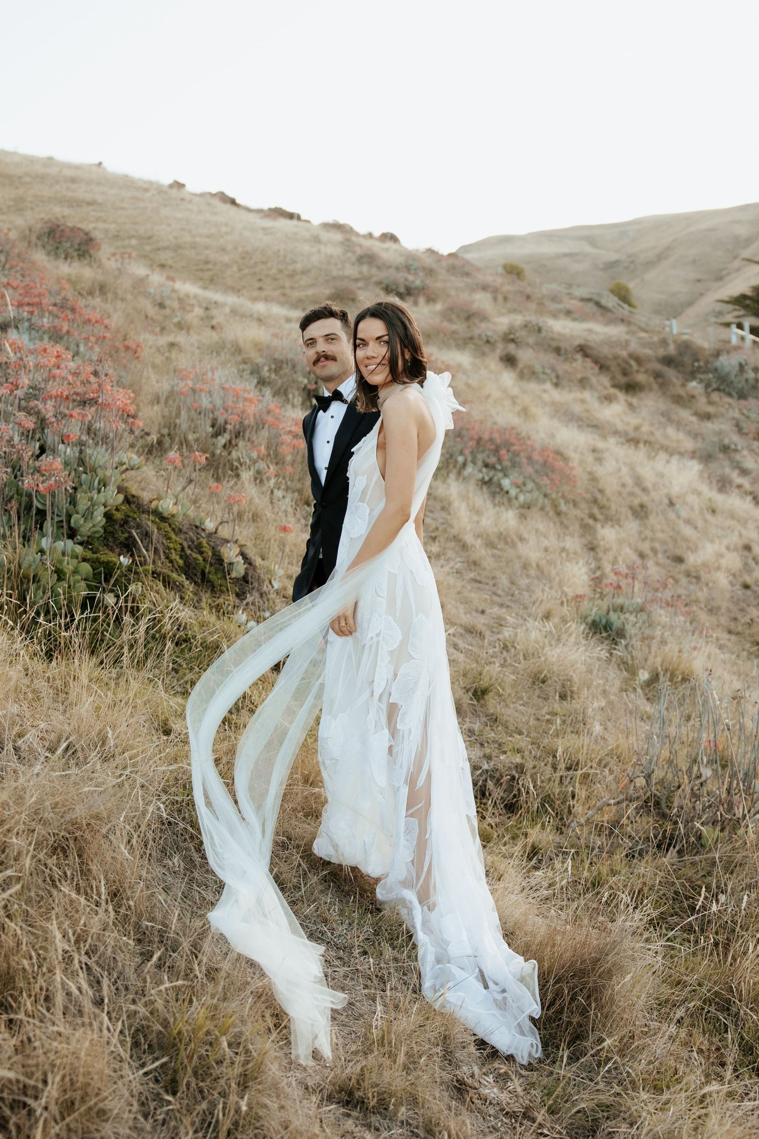 Bride's white lace dress flows in wind while taking romantic hillside portraits.