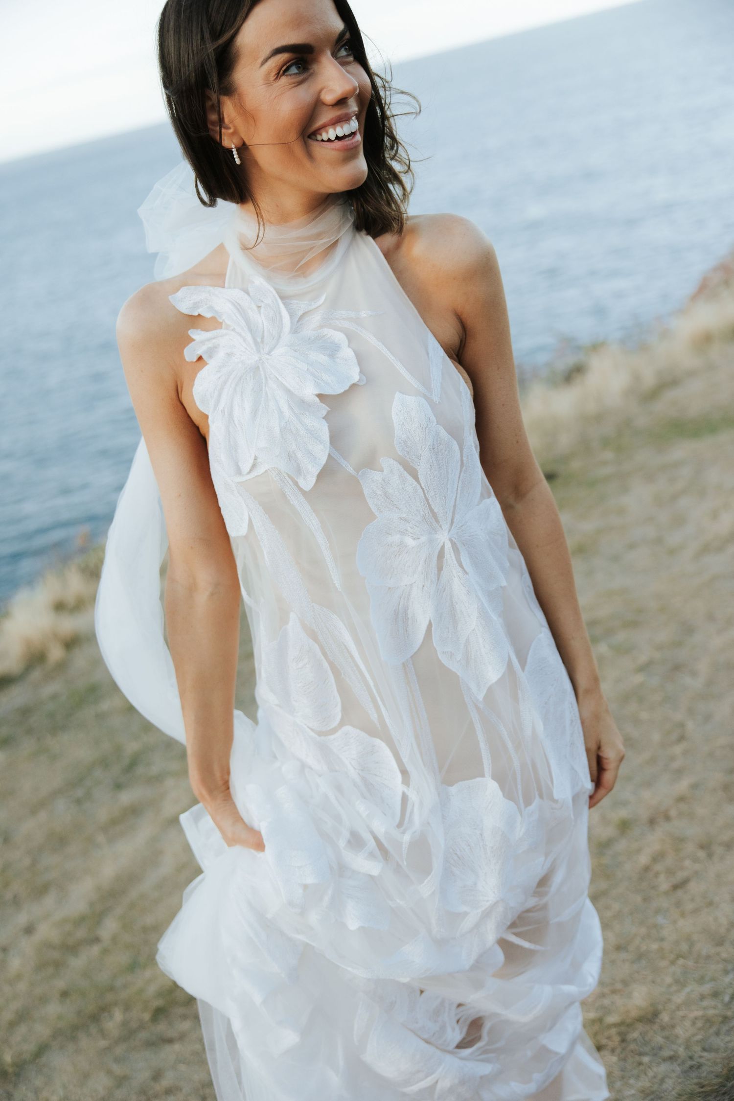 A bride wears a stunning white halter dress with floral appliques against an ocean backdrop.