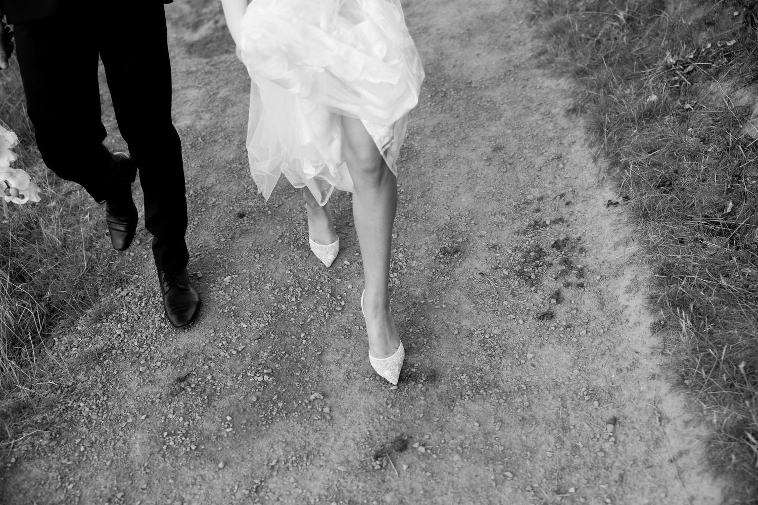 Black and white photo showing wedding dress and suit-clad feet walking along a dirt path.