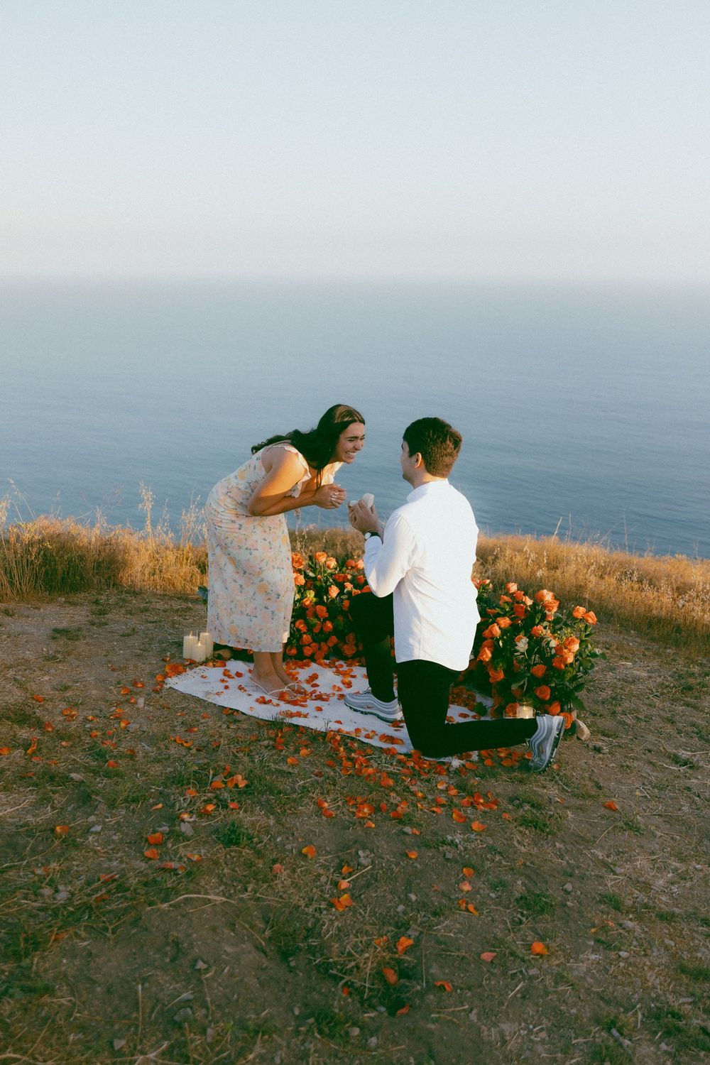 A romantic sunset proposal setup on a coastal cliff with orange flowers scattered on a blanket overlooking the ocean.