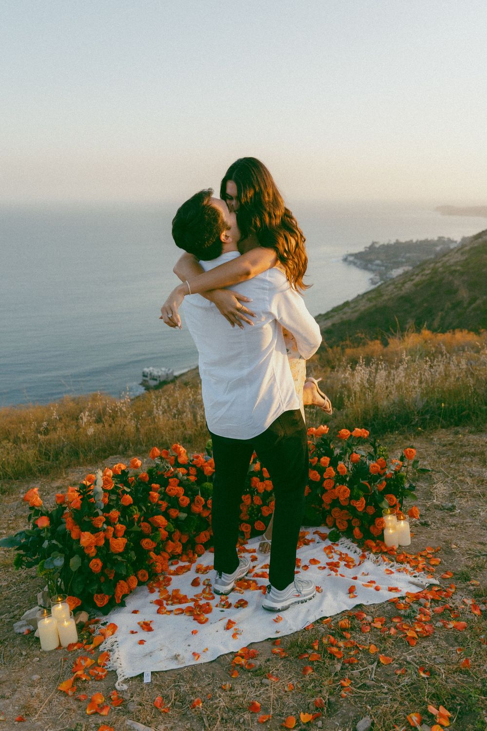 A romantic couple embraces on a scenic coastal cliff surrounded by orange California poppies and candles at sunset.
