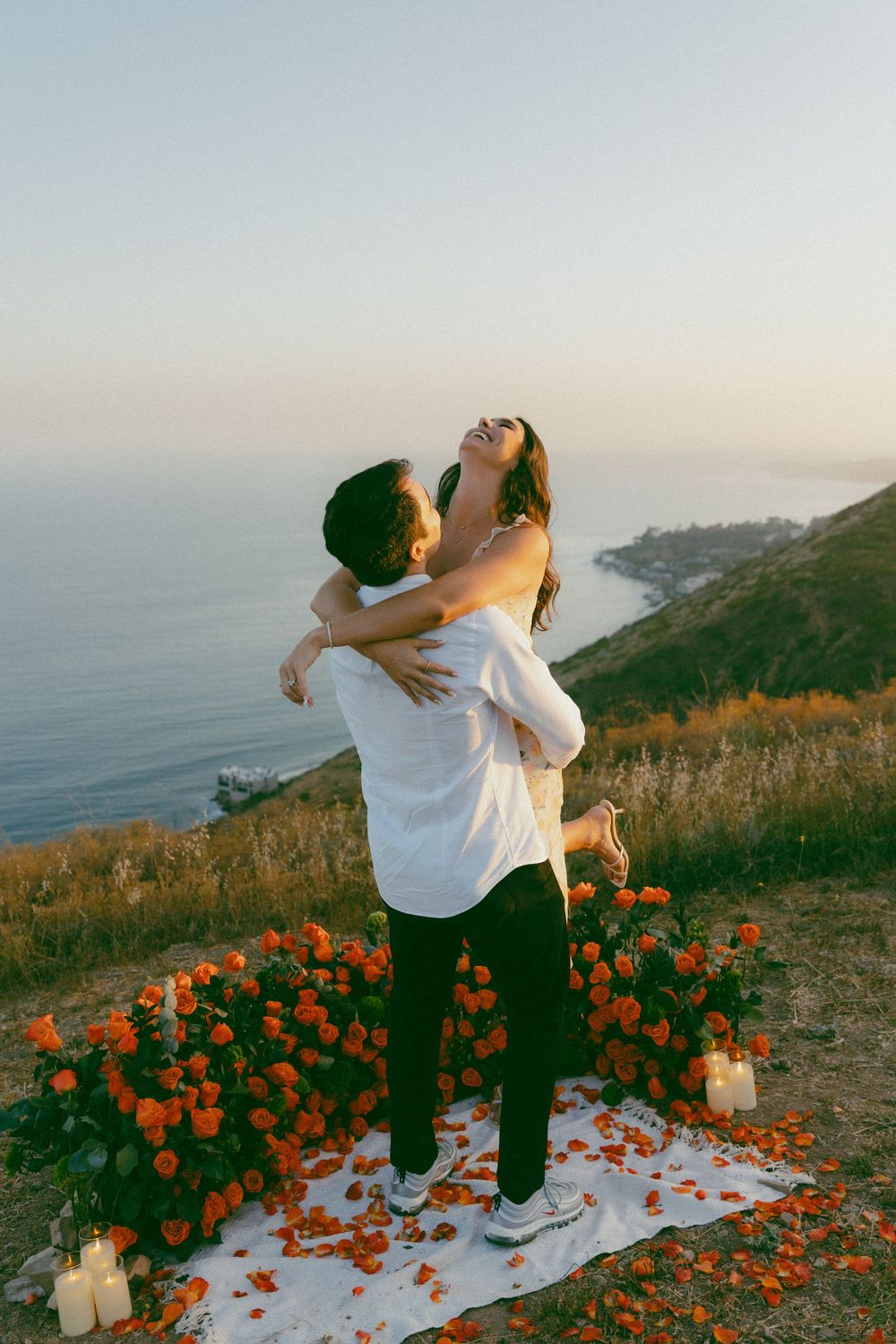 Romantic couple embraces on coastal clifftop at sunset surrounded by orange flowers and candles overlooking the ocean.