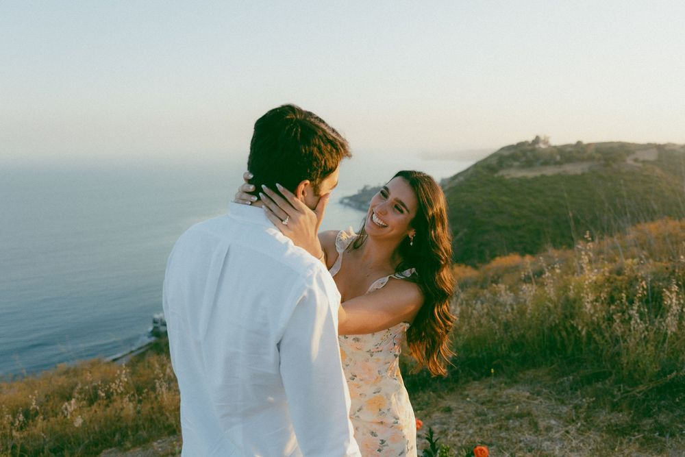 A romantic couple in white embraces on a scenic coastal cliff during sunset.