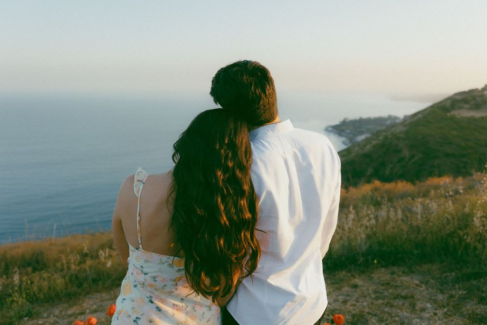A couple sits together overlooking a coastal view from a grassy cliff during golden hour.