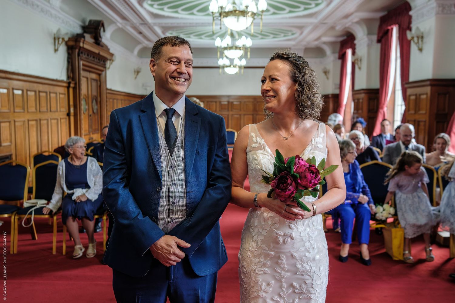 A joyful couple shares a laugh during their wedding ceremony in an elegant wood-paneled room with red curtains.