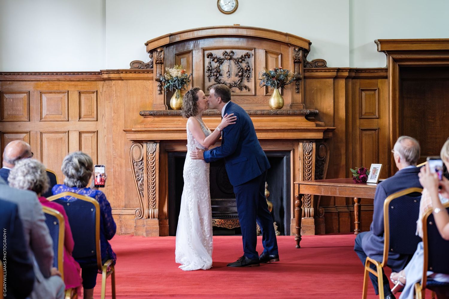 A wedding ceremony takes place in a traditional wood-paneled chapel with ornate details and guests seated in pews.