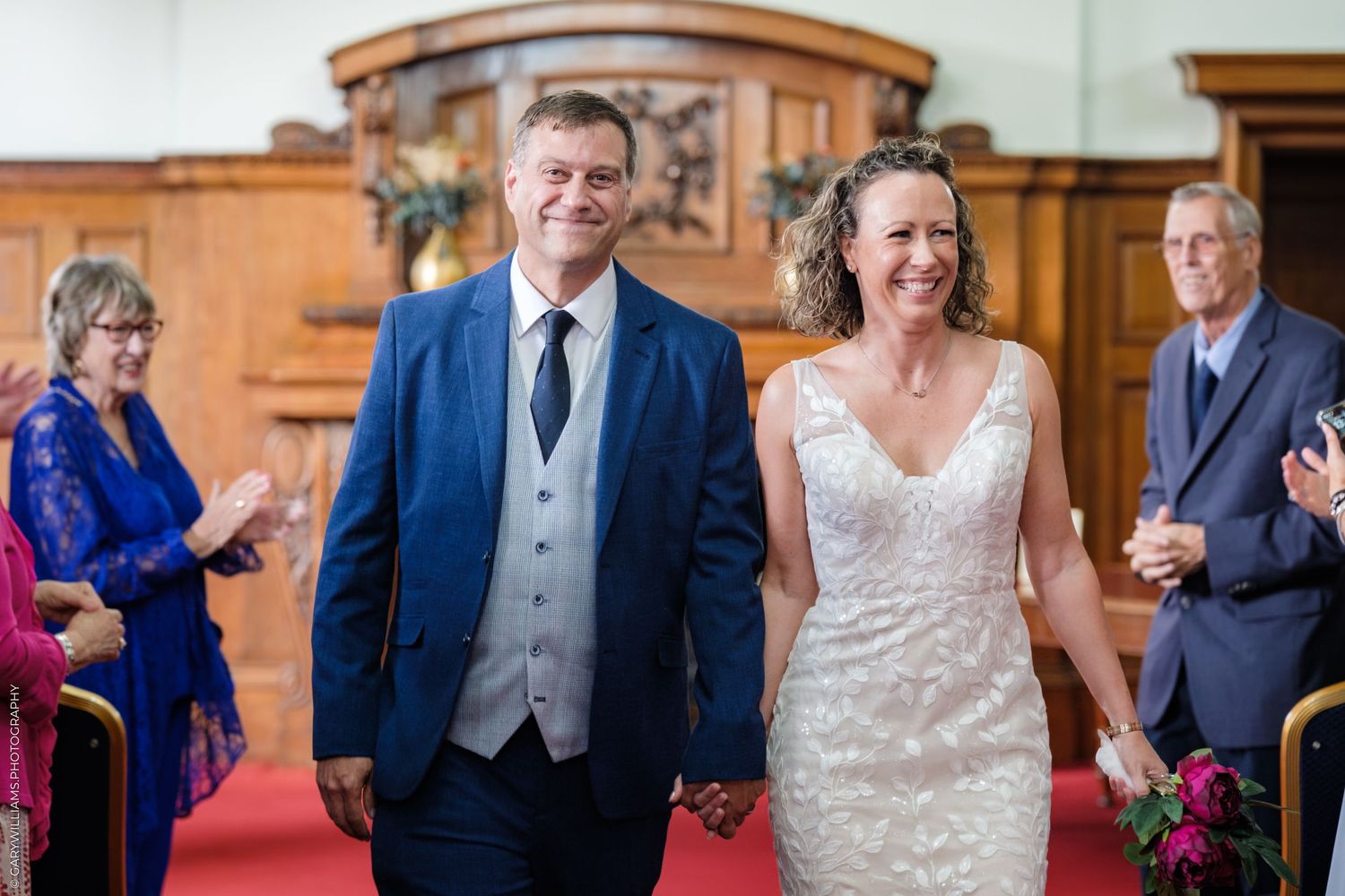 A couple holds hands during their wedding ceremony in an elegant wood-paneled room with red carpet and onlookers present.