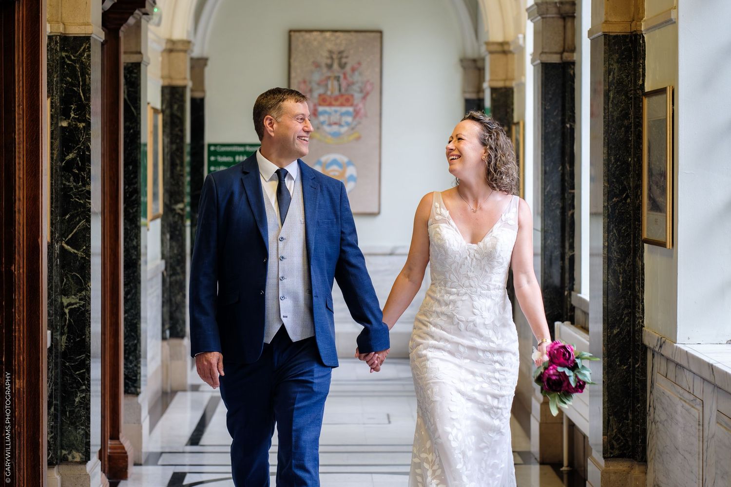 Two people walk hand in hand down an elegant hallway with marble columns while smiling at each other on their special day.