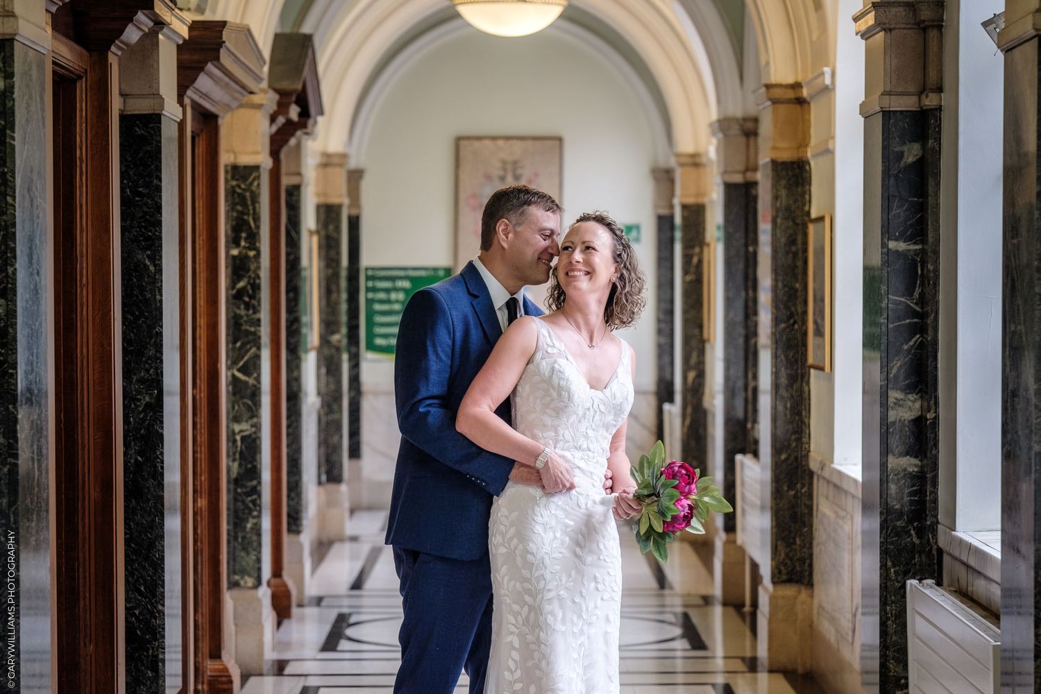 A happy couple in wedding attire laughs together in an elegant marble-columned hallway with arched ceilings.