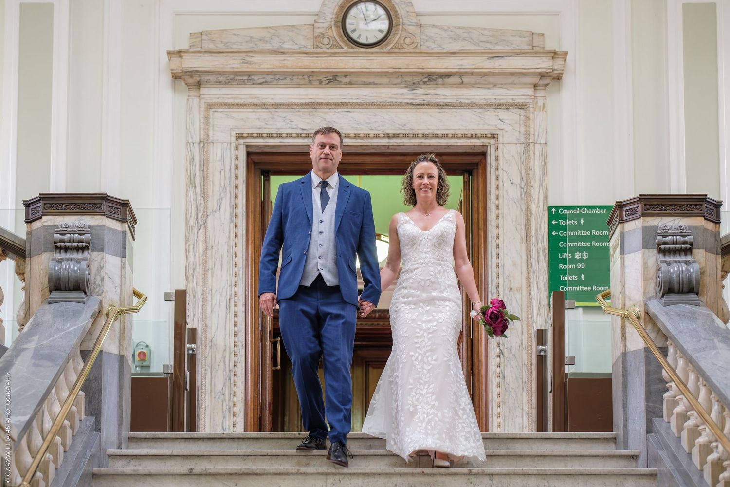 A bride and groom walking through an elegant marble hallway with ornate architectural details and a wall clock mounted above.