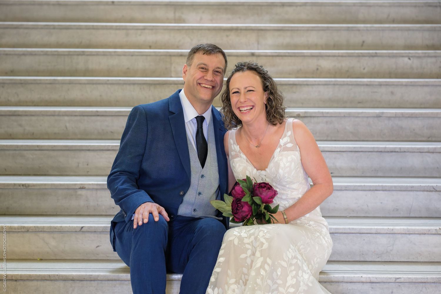 A happy couple sits on stone steps on their wedding day, with the bride holding a deep red and pink bouquet.