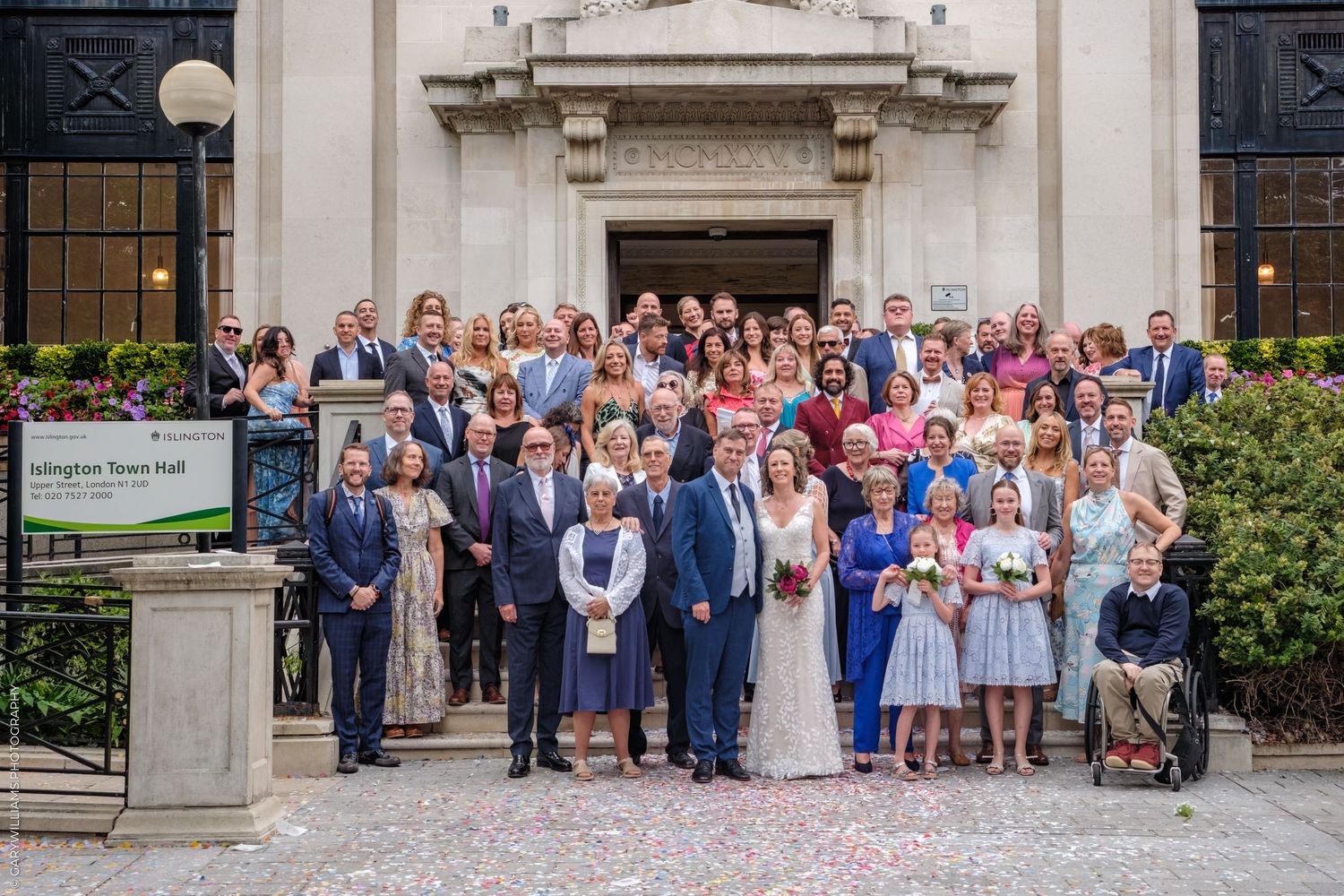 Large wedding party gathered on stone steps outside historic building entrance.