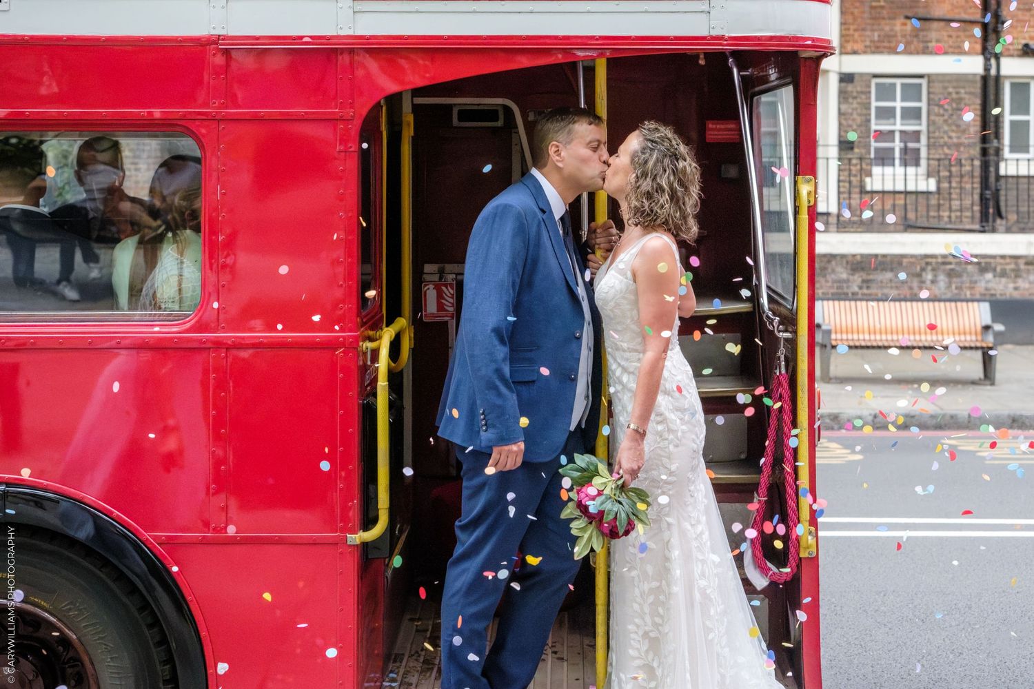 A romantic wedding moment captured in front of a bright red vintage London bus with confetti falling.