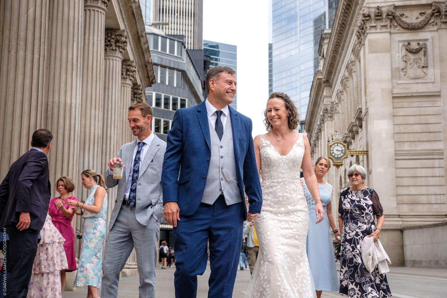 A celebratory wedding scene with classical architecture and city buildings in the background.