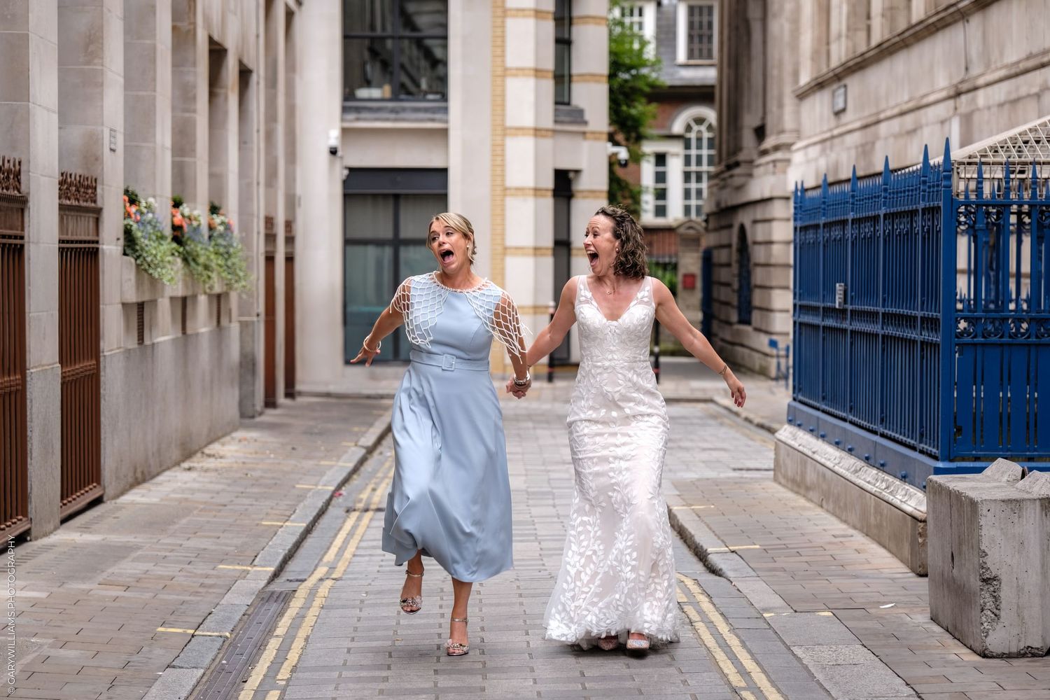 Two people walk along an urban alley, one wearing a blue dress and one wearing a white lace gown.