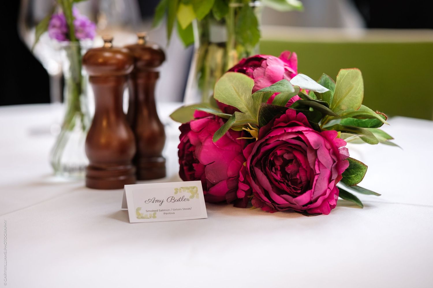 Deep red roses with green leaves rest on a white tablecloth next to a place card and wooden salt shaker.