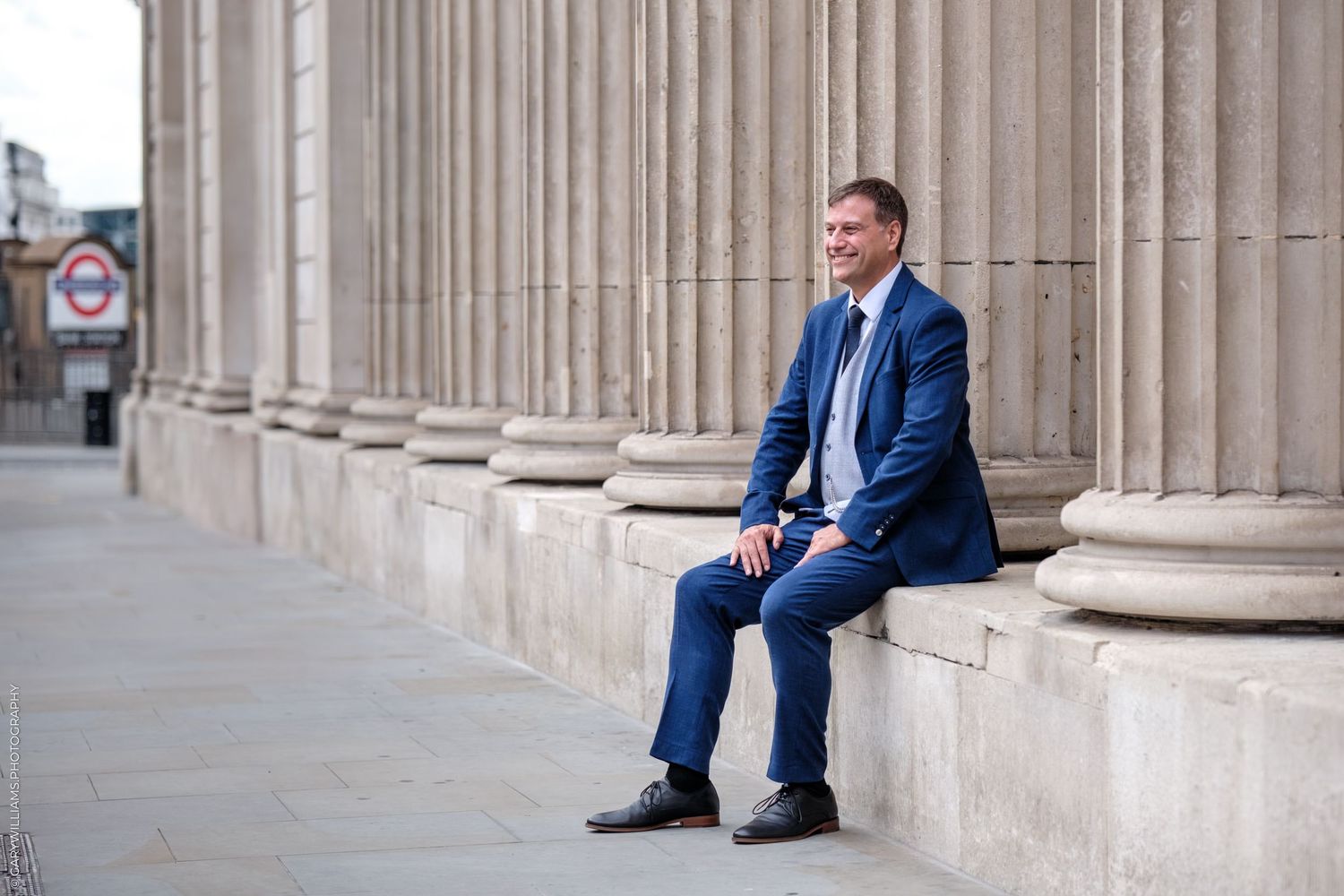 Professional in a navy blue suit sits on stone steps between classic white columns of a grand building.