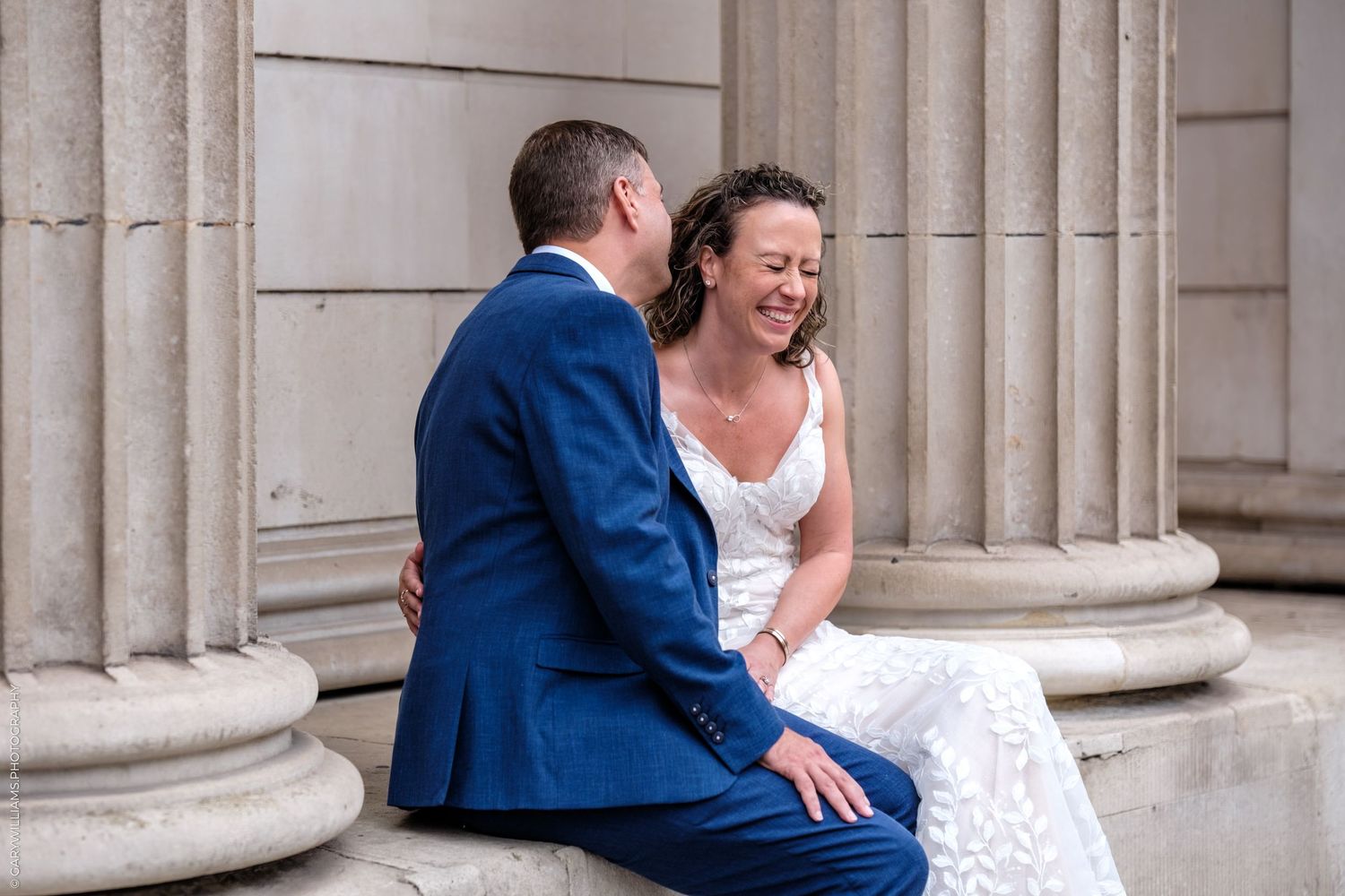 A couple in formal wedding attire shares an intimate moment among classical stone columns.